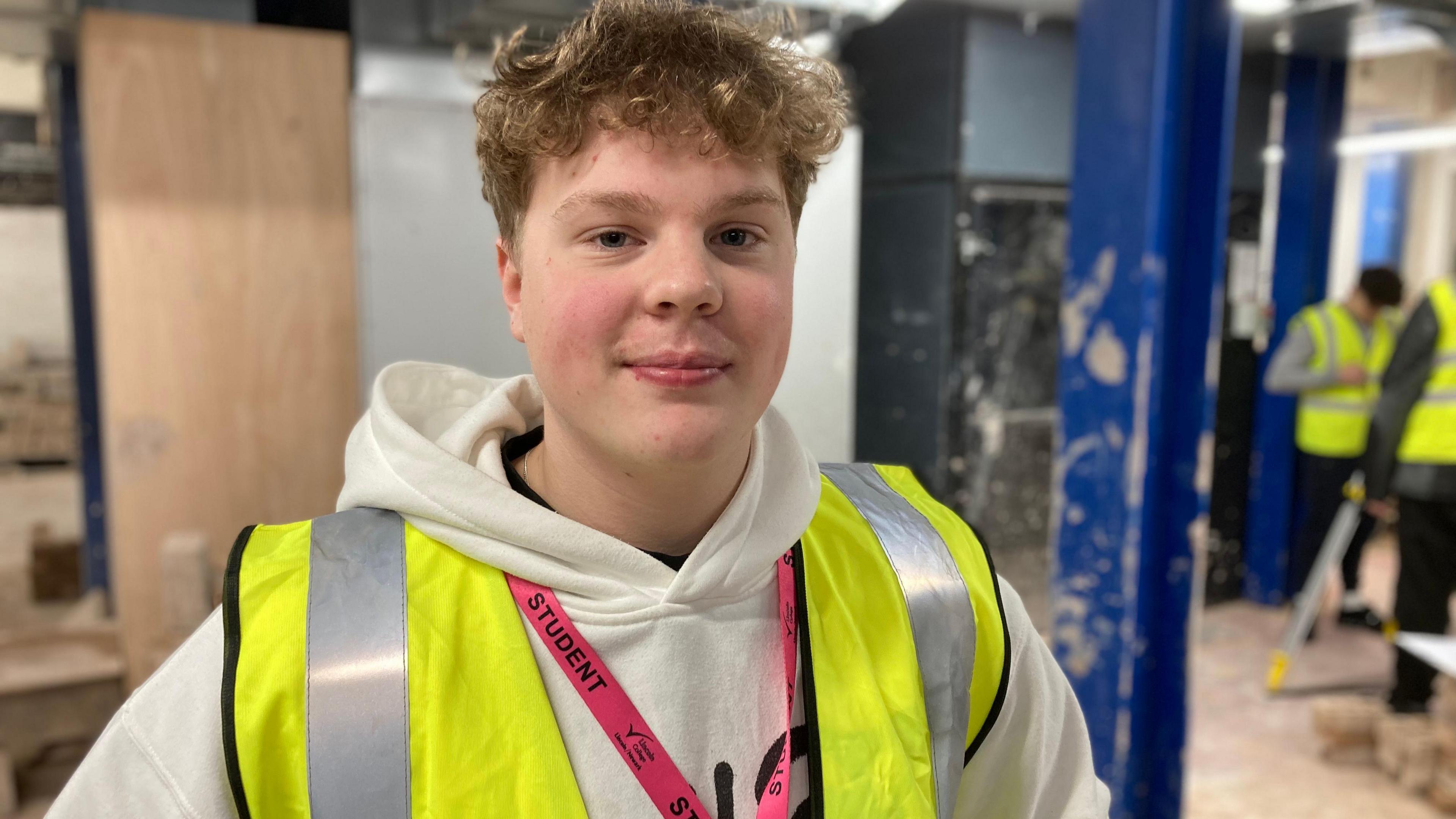 A teenage boy with light brown curly hair is standing in a construction environment. He is wearing a high-vis jacket over a white hooded top and in the background are other students and tools.