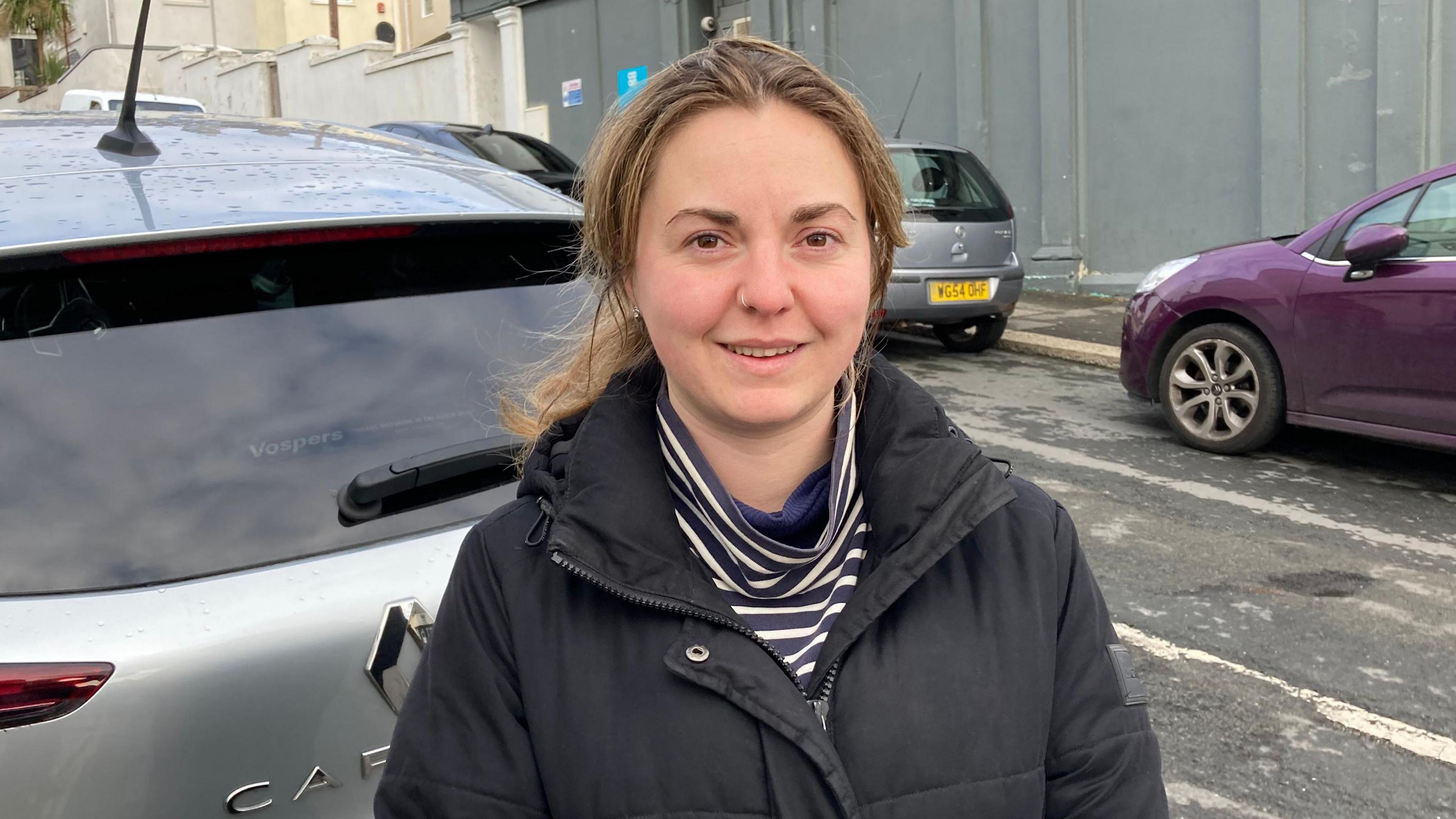 Katherine Terris wearing a black coat and a blue and white stripey top stands in front of a silver Renault car. There are purple and silver cars on the opposite side of the road. Katherine had a nose piercing.