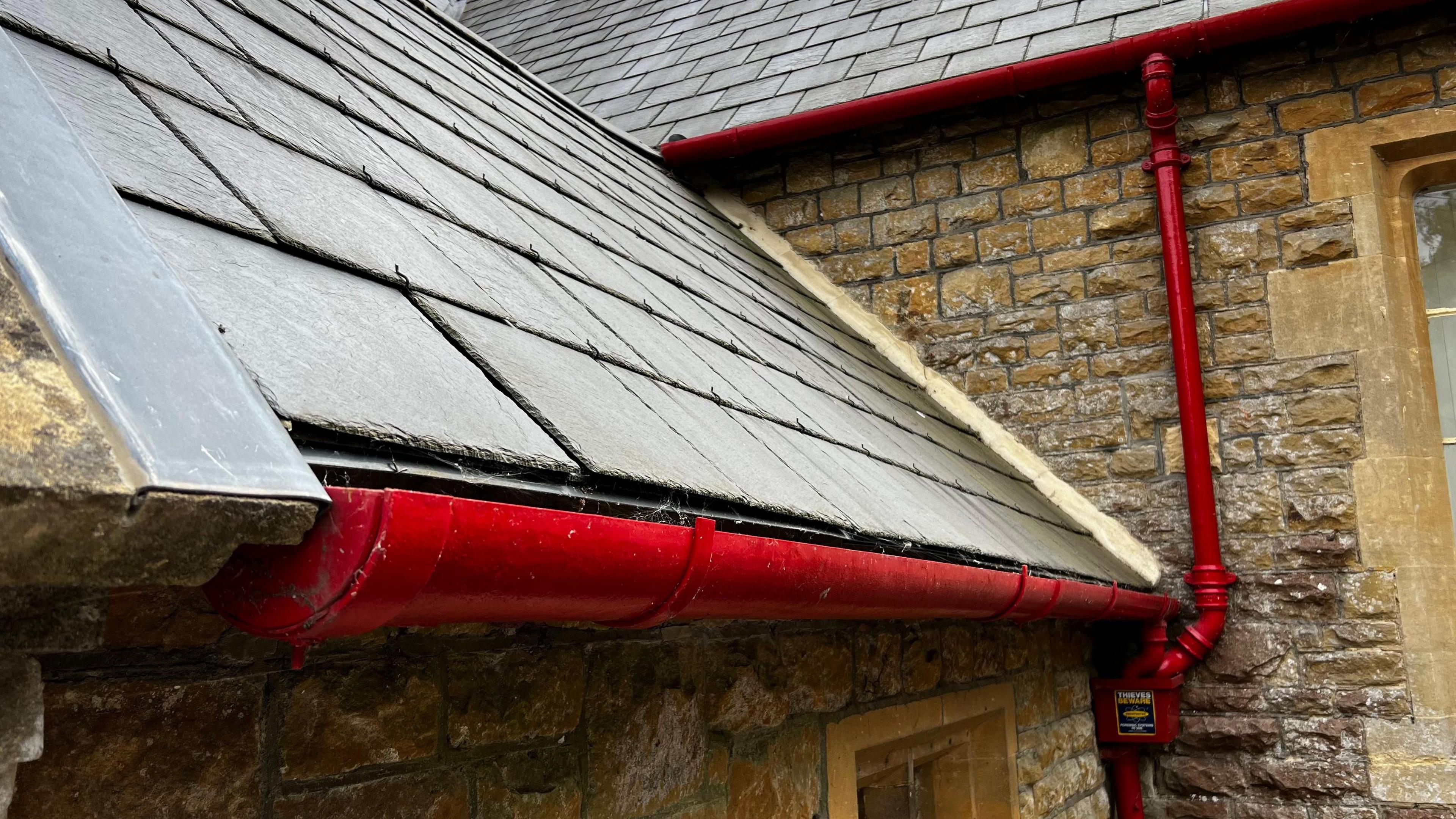 The outside of a stone with slate-like tiles on its roof and bright red iron gutting running along the edge of the roof.
