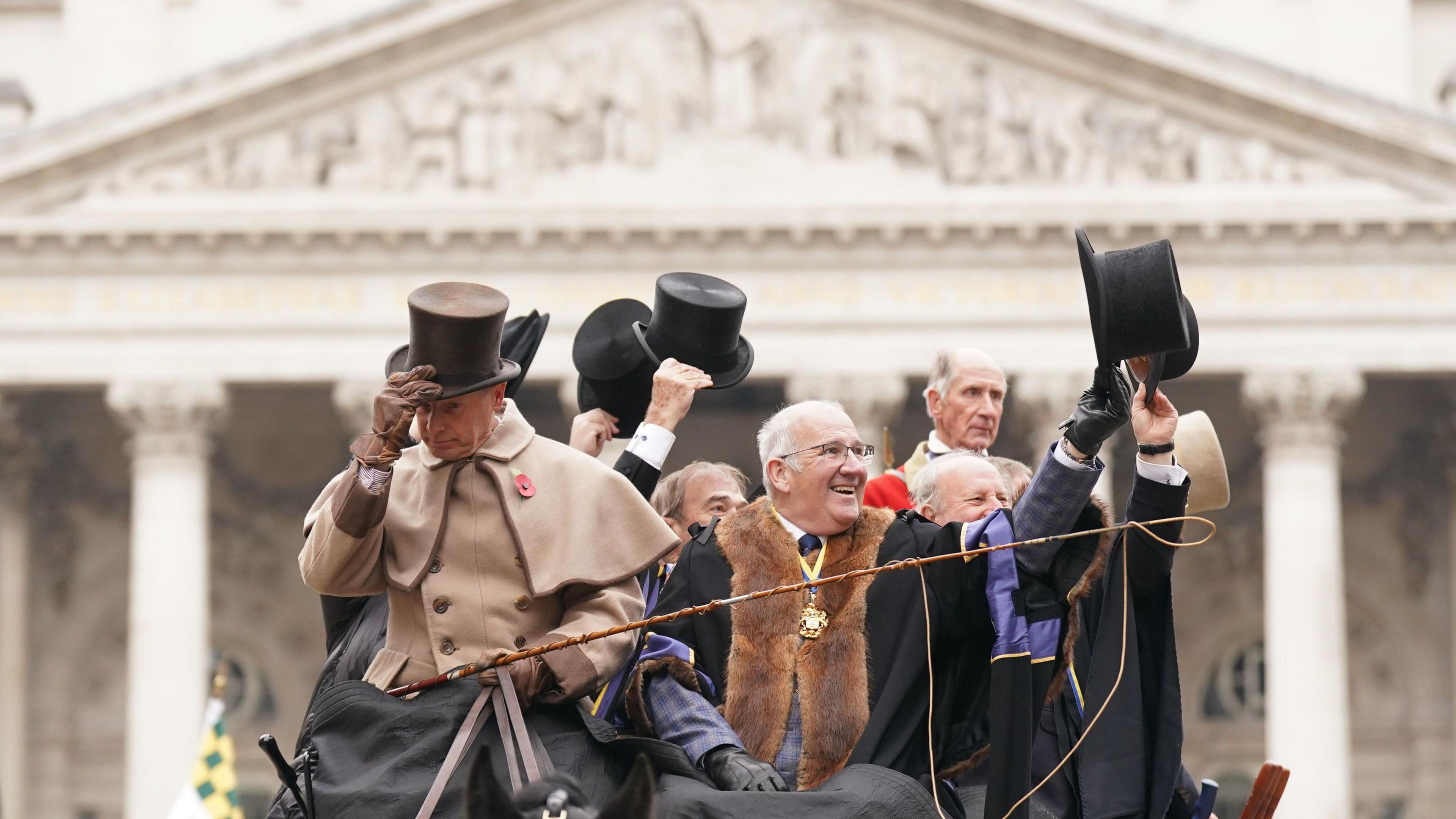 A group of men dressed in traditional attire, including top hats and ceremonial robes. They wave and tip their hats to the crowd from a horse-drawn carriage, with a grand building featuring classical columns and carvings in the background.