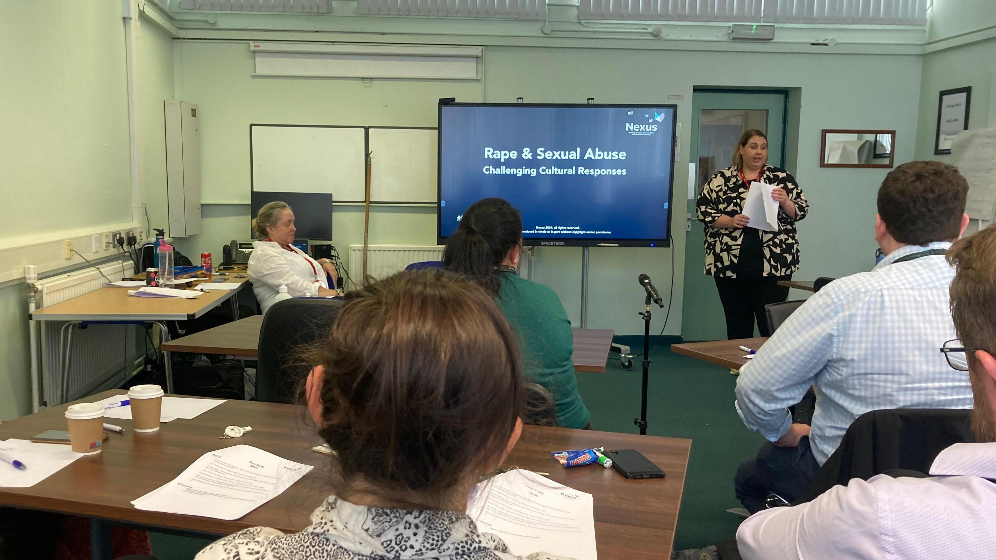 A room full of people, most with their backs towards the camera. Jan Winton, from Nexus, is standing at the front of the class beside a screen which has the words "Rape and Sexual Abuse, Challenging Cultural Responses" written on it