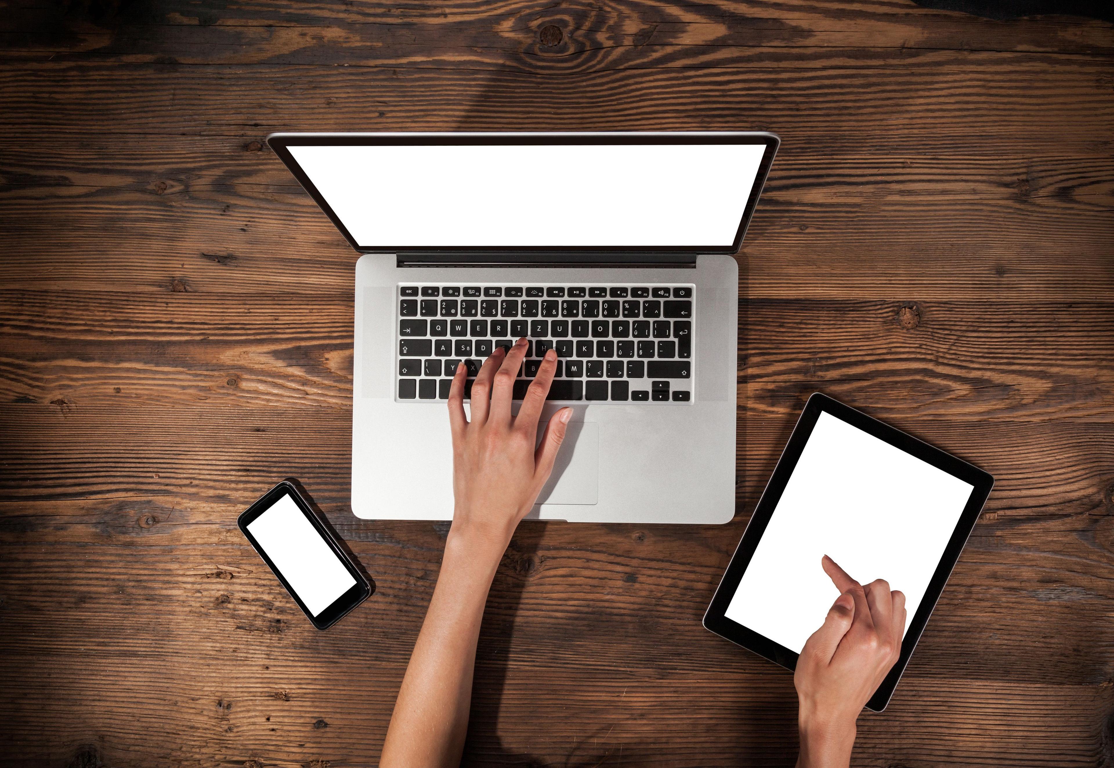 A laptop, a phone and a tablet are on a wooden table. Two white hands are shown typing and working on the tablet. 