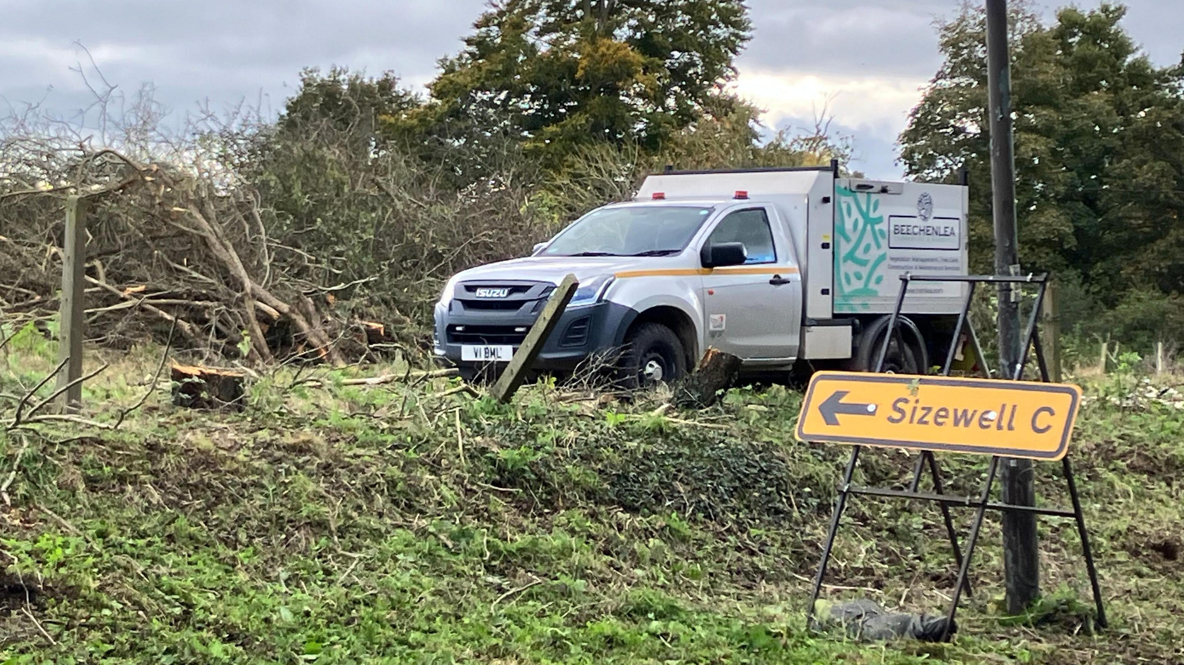 A white van is parked on an area of land where vegetation has been cleared in front of a yellow sign reading "Sizewell C" with a black arrow on it  