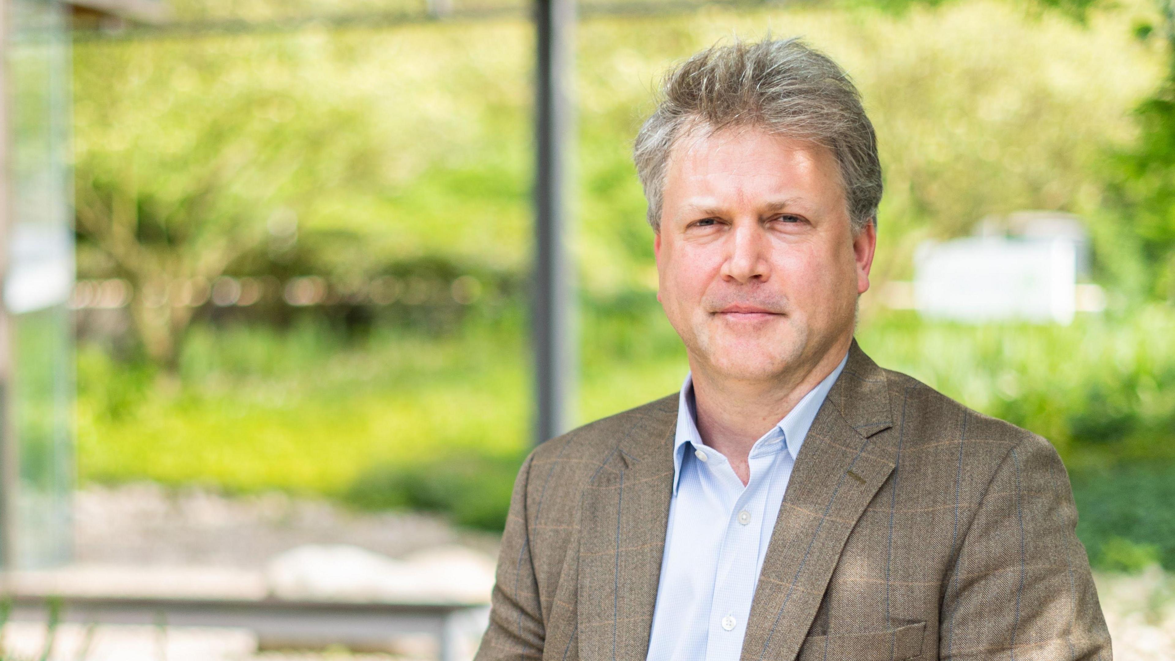 The male professor, who has greyish-brown hair, stares at the camera wearing a blue shirt and brown checked suit - there is a blurred green background of trees and bushes behind hime