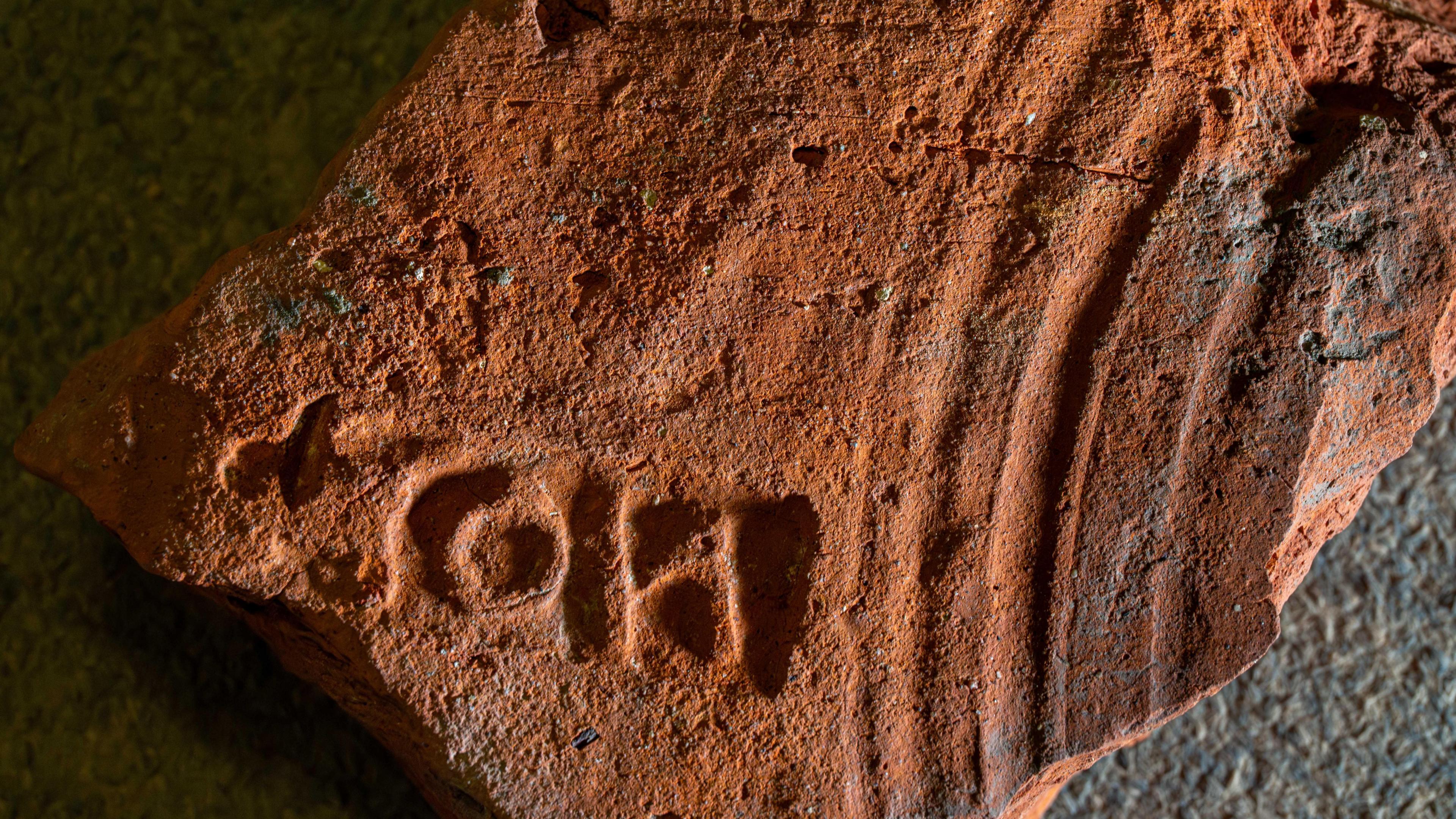 A red roof tile has a stamp made up of three letters on its surface showing that it was a public building and belonged to the procurator of London. It also has a series of curved grooves made by the tile maker dragging his fingers across it when it was being produced. 