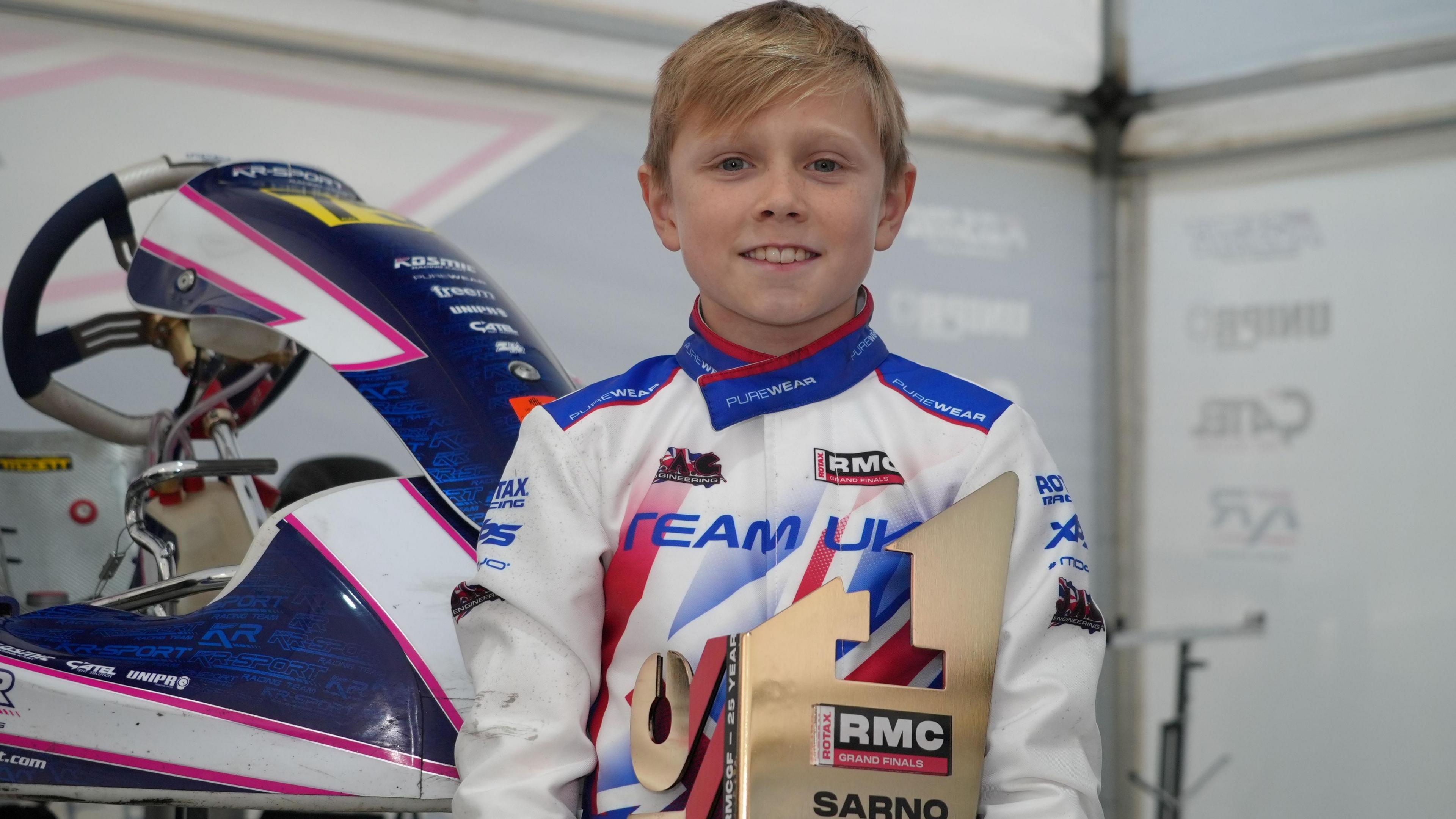 Josh Cooke in his white race suit with sponsors' logos, posing with his Rotax World Championship trophy. He is smiling. A kart can be seen behind him.