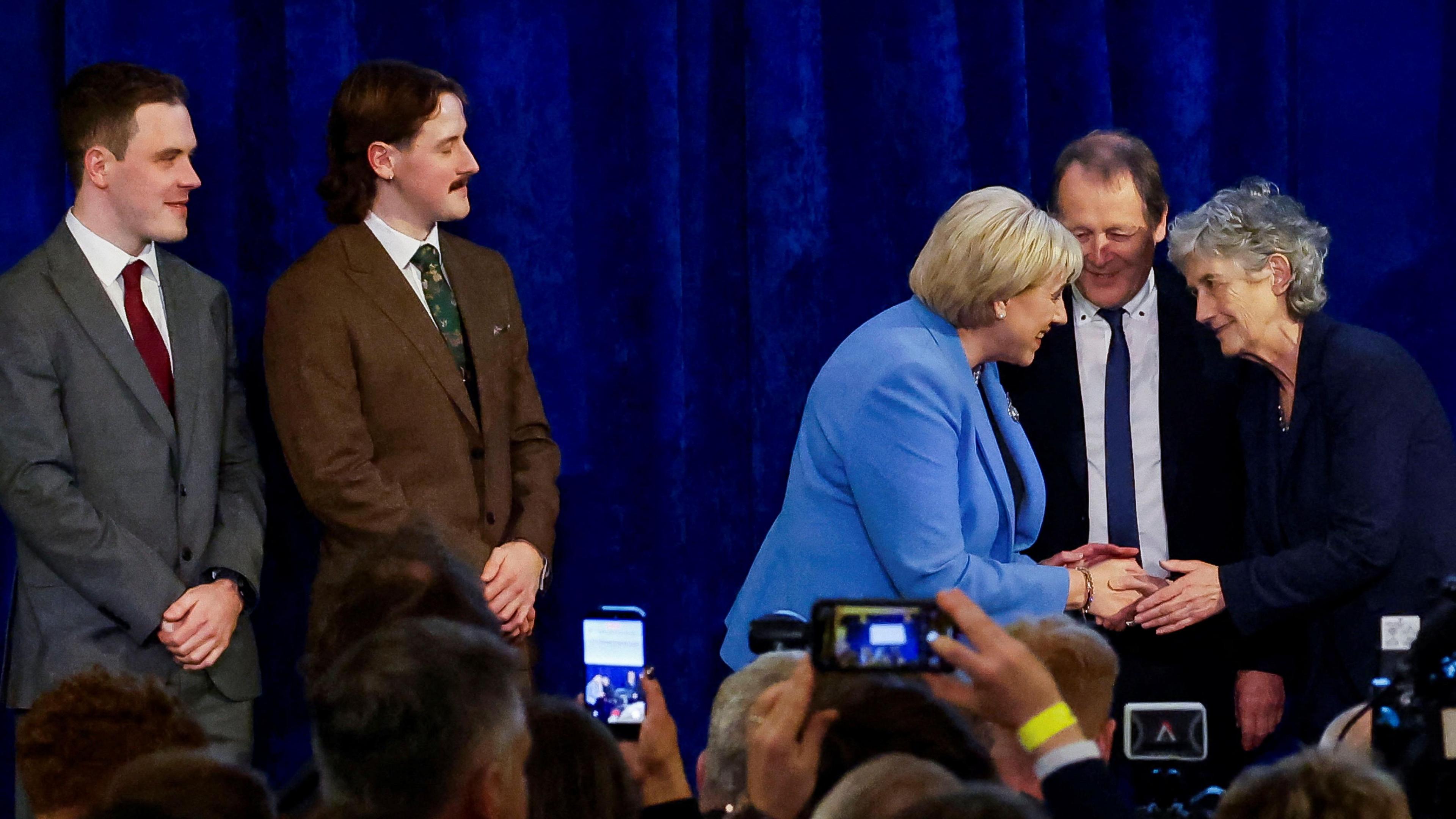 Two women shake hands on stage. The woman to the left has short blonde hair and is wearing a blue blazer, the woman two the right has short hair and is wearing a navy blazer. A man in a suit and blue tie stands behind them. Two other men is suits stand to the left and people in the crowd are taking pictures with their phones.