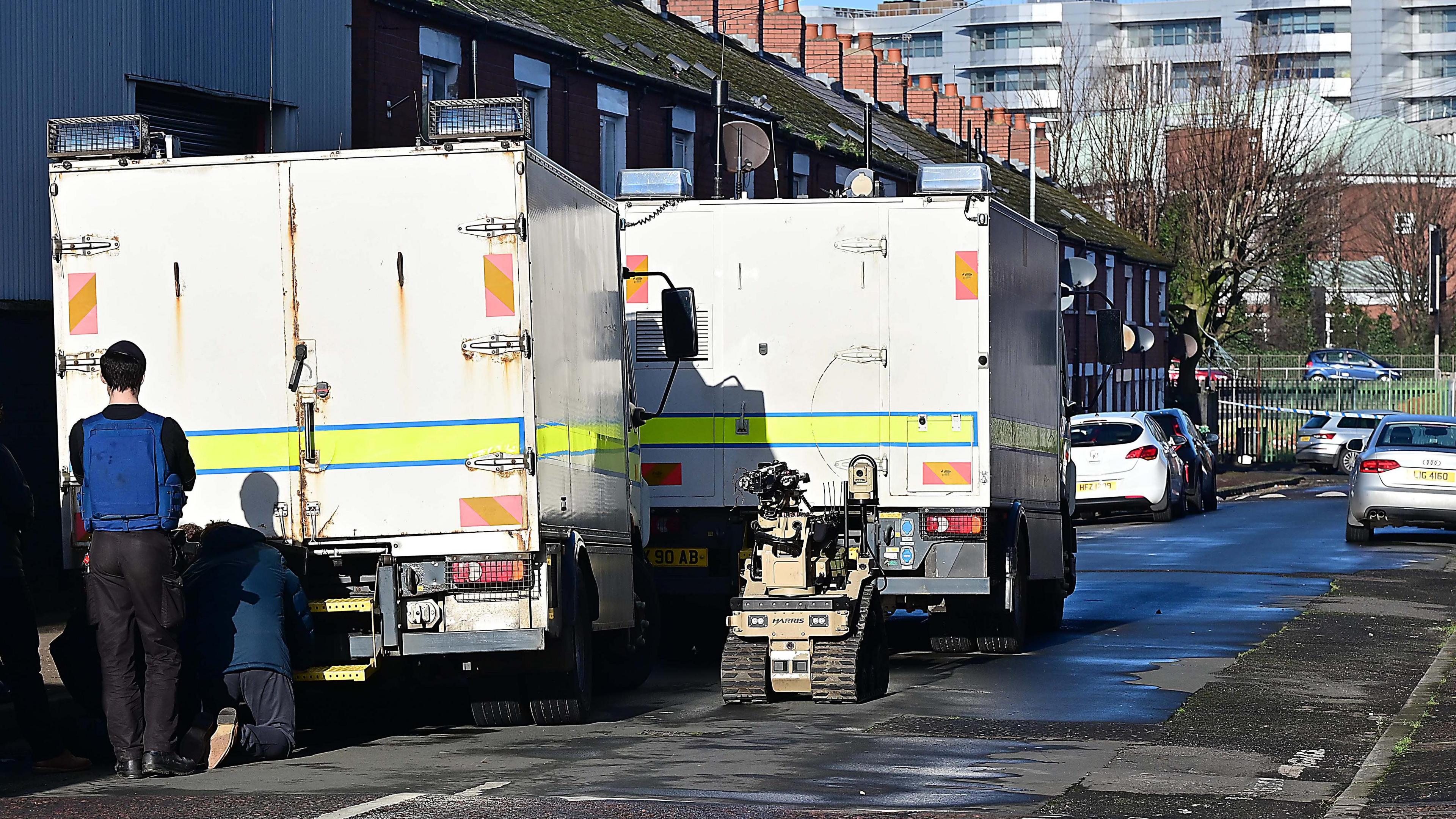 Police and Army Technical Officers stand by a white van with what looks like a bomb disposal robot. The street is in view with three cars parked outside houses. 