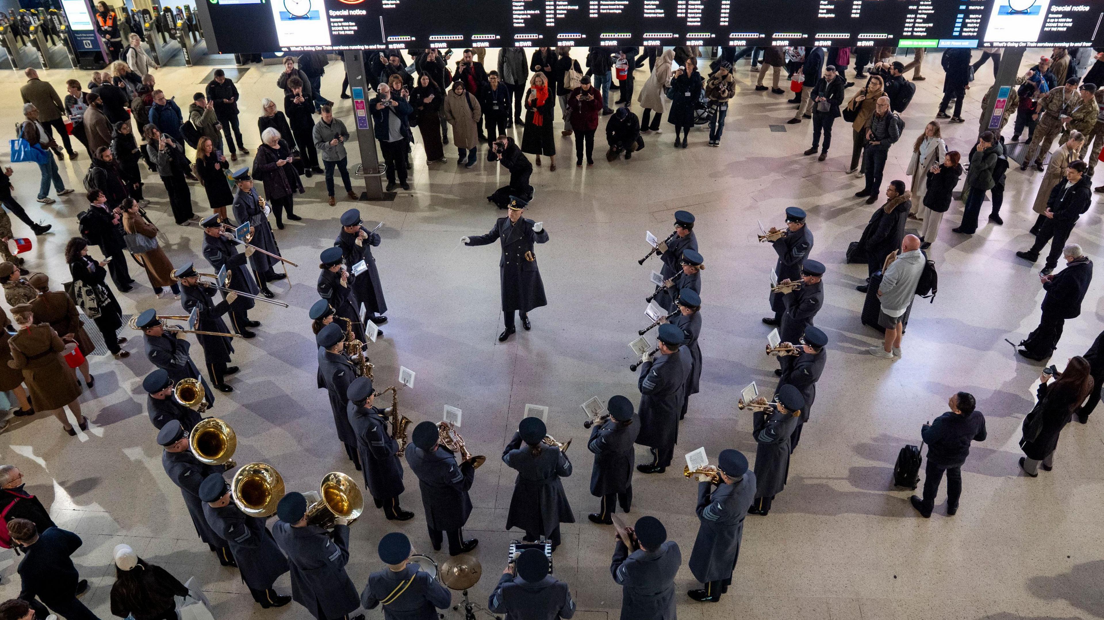An aerial shot shows the RAF Band standing in a horseshoe of two ranks of musicians. Their conductor is facing the camera and the musicians have their backs to the camera. They are wearing overcoats and hats and many of them are holding gold coloured brass instruments. A digital timetable is behind them and  commuters are standing around them.