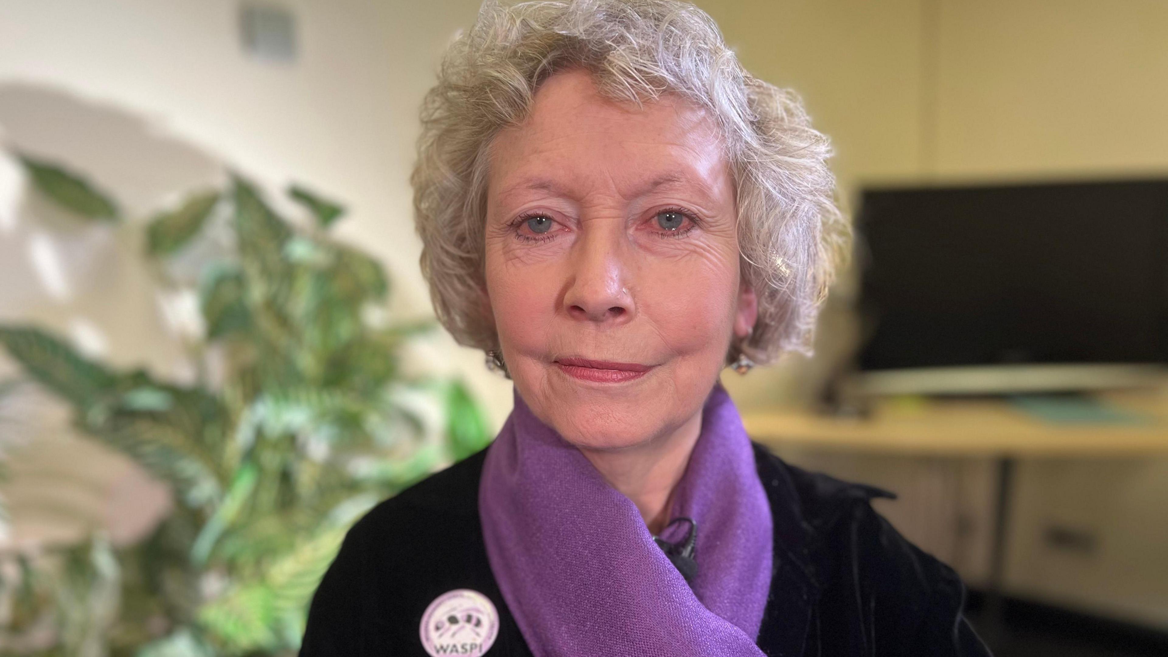 A woman in her 70s looking pensive with a purple scarf and a WASPI badge. 