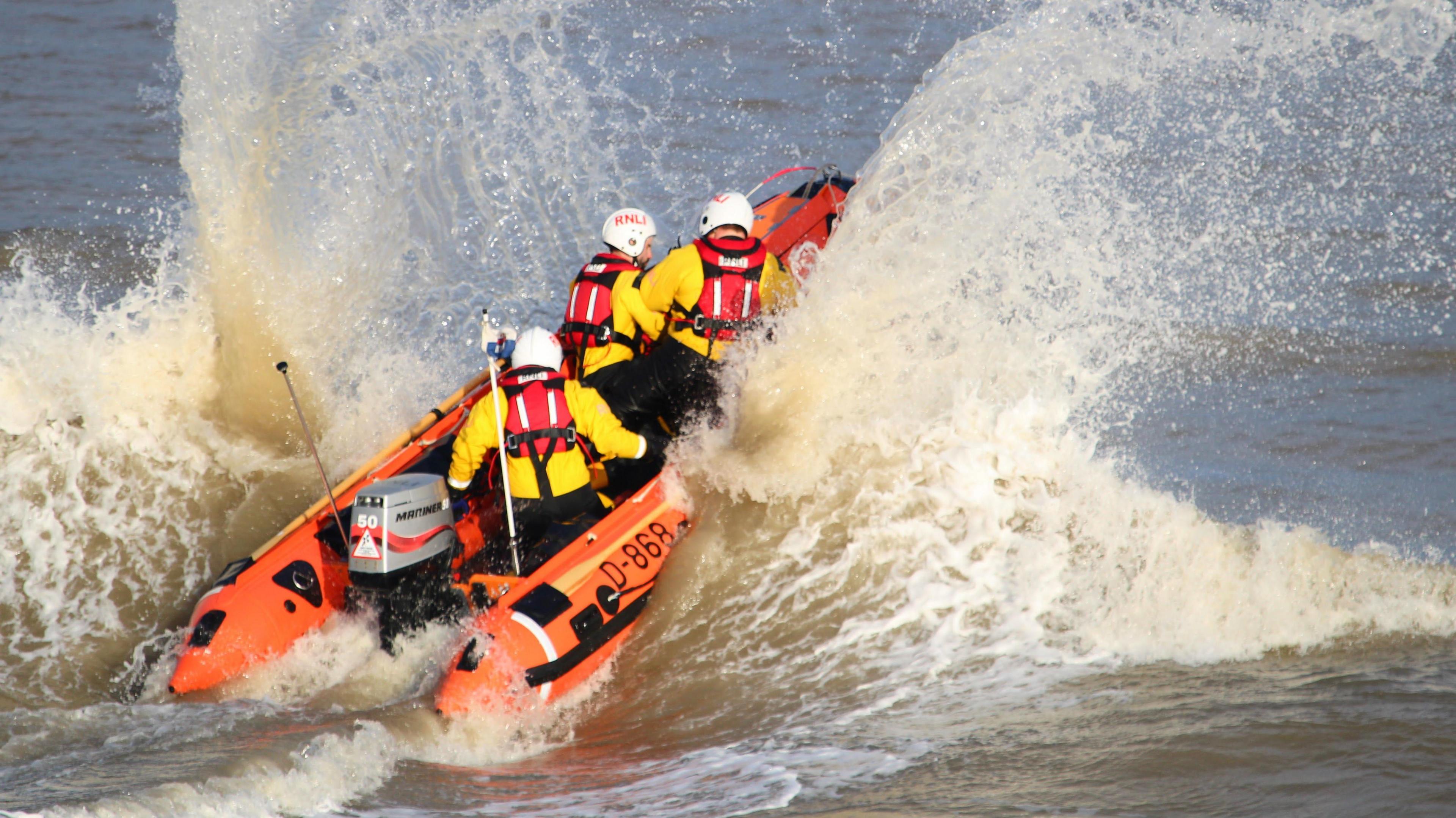 A small orange boat, with three crew members wearing bright yellow coats and red life jackets, bounces on the sea with spray splashing up against the sides of the boat and in its wake.