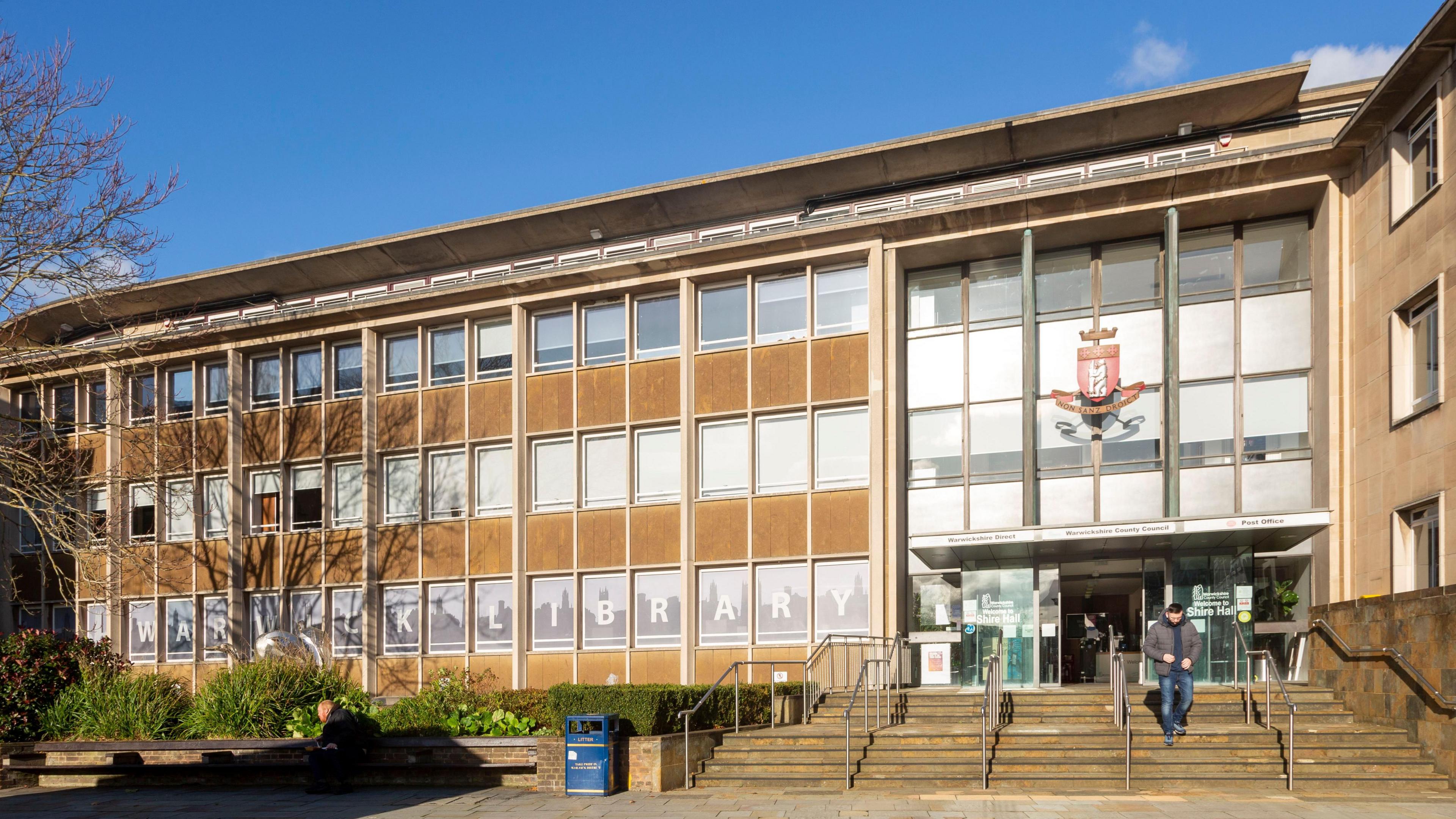 The County Council building in Warwick. It has lots of small square windows and large windows above the entrance. There is the council logo above the doors. There are steps with hand railings leading up to the entrance