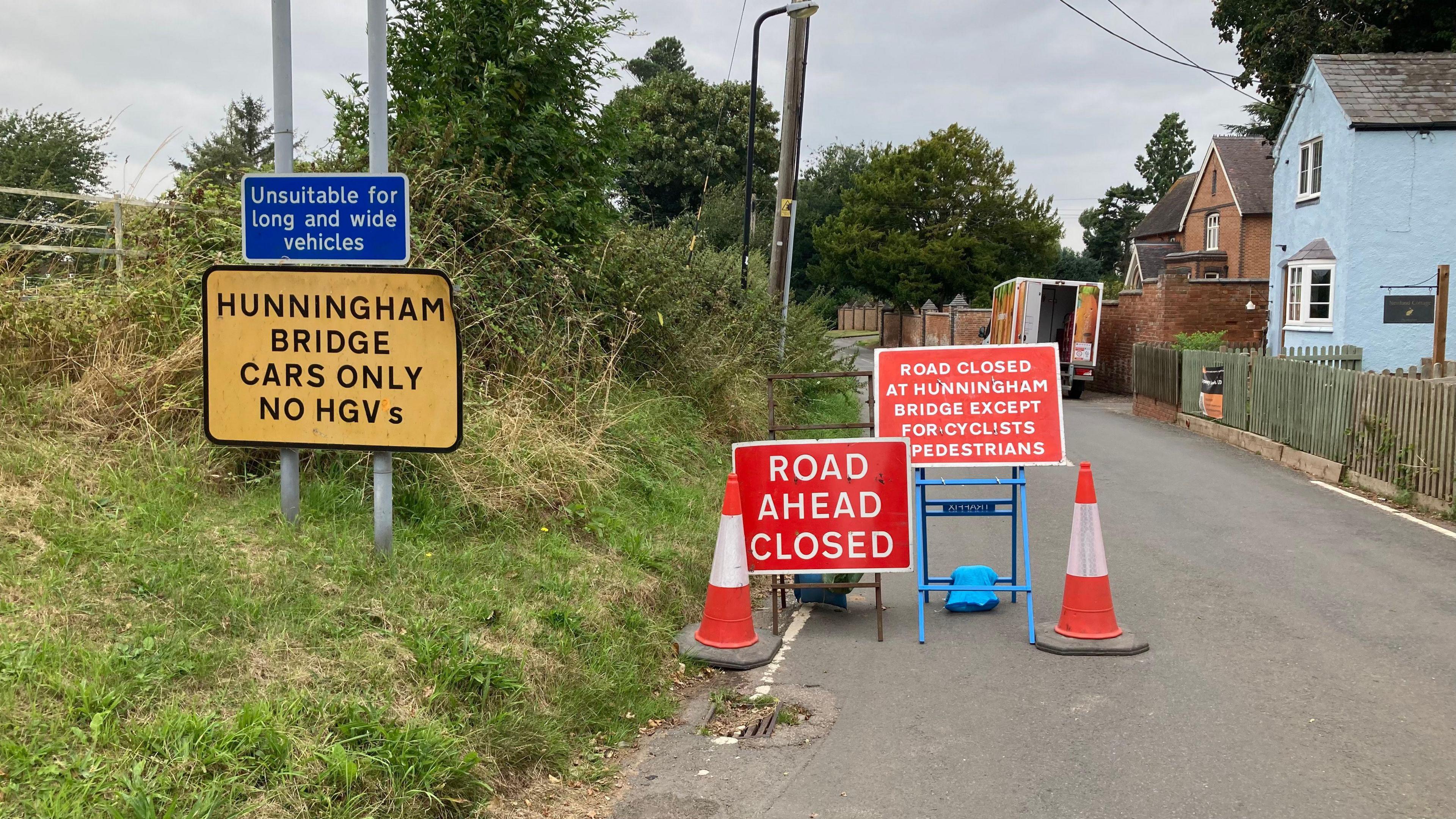 Two cones and red temporary road closure signs in the middle of the road, with a yellow permanent sign attached to a signpost saying the bridge is only suitable for cars and not HGVs.