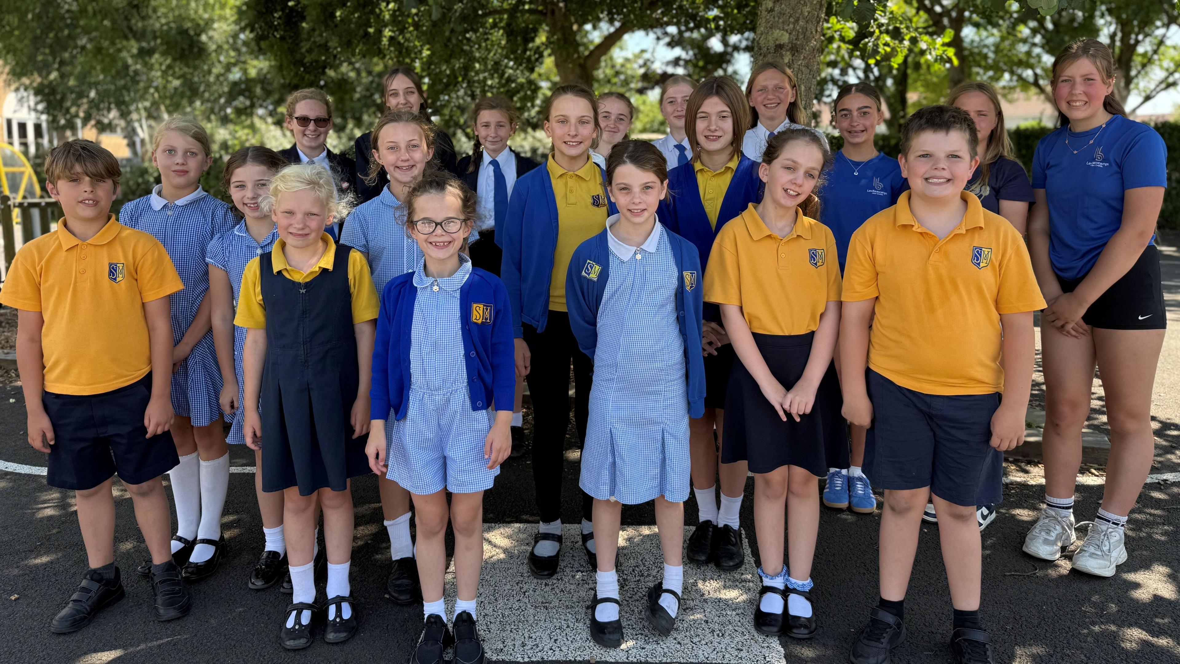 A group of school children in three rows, wearing school uniform in black and yellow, and blue gingham stand together smiling at the camera.