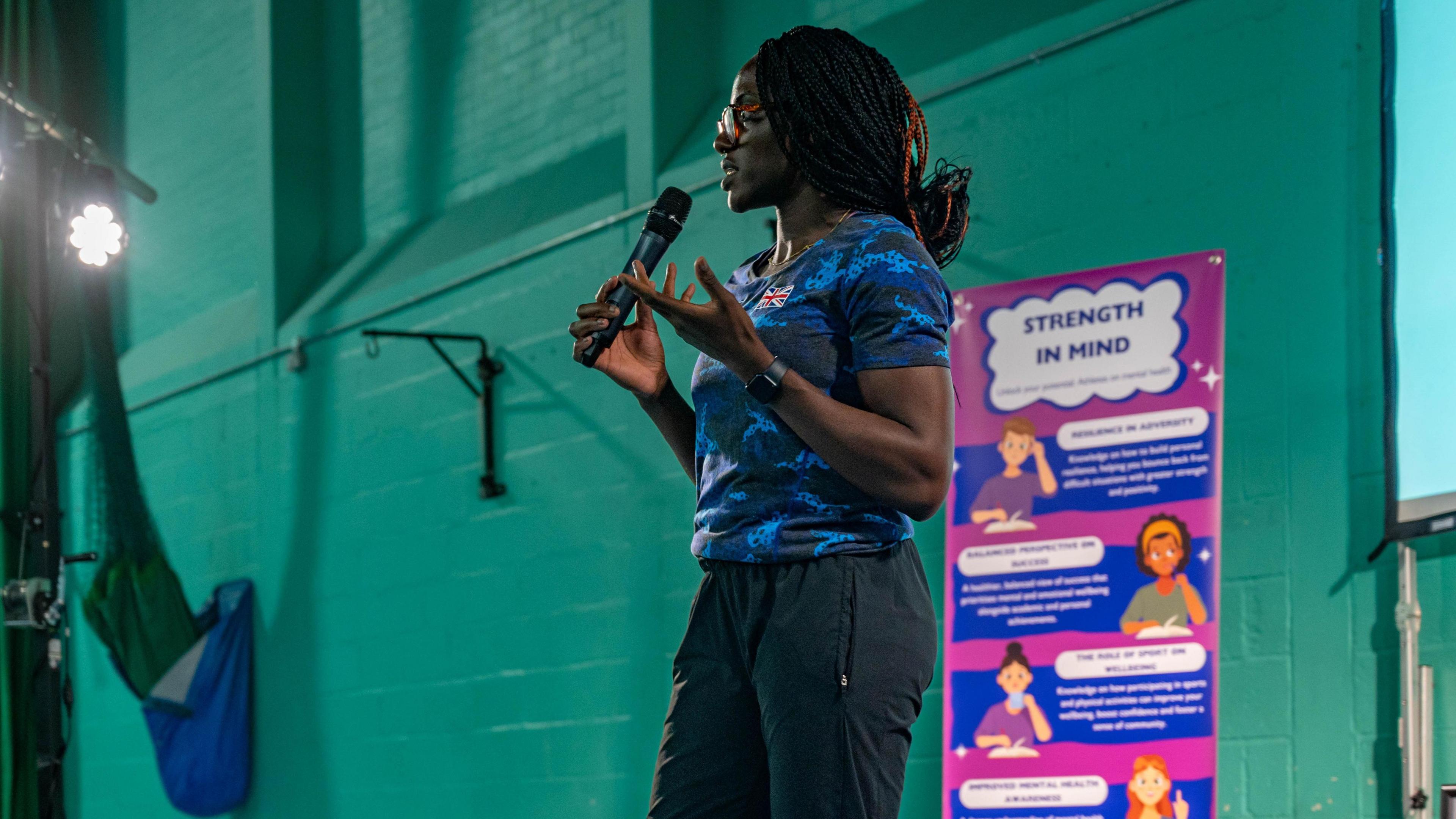 Olympic sprinter Victoria Ohuruogu is on stage at the event, holding a microphone, The wall behind her is painted green bricks, and in front of her are several rows of children, listening to her speak.