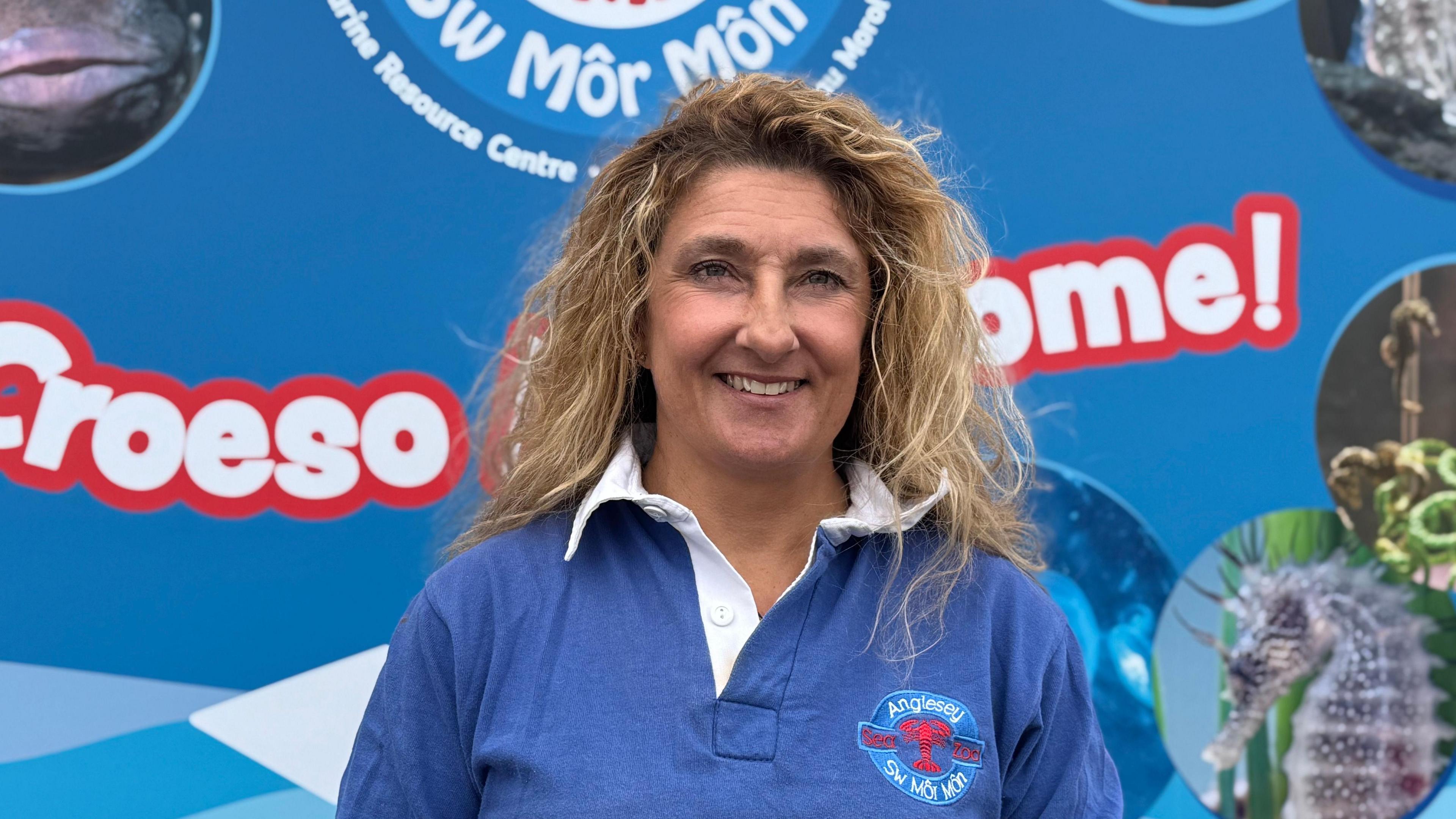 Frankie Hobro, a woman in her early 50s with blonde hair, looking at the camera. She's wearing a blue polo shirt with the logo of the Anglesey Sea Zoo and standing in front of an Anglesey Sea Zoo banner