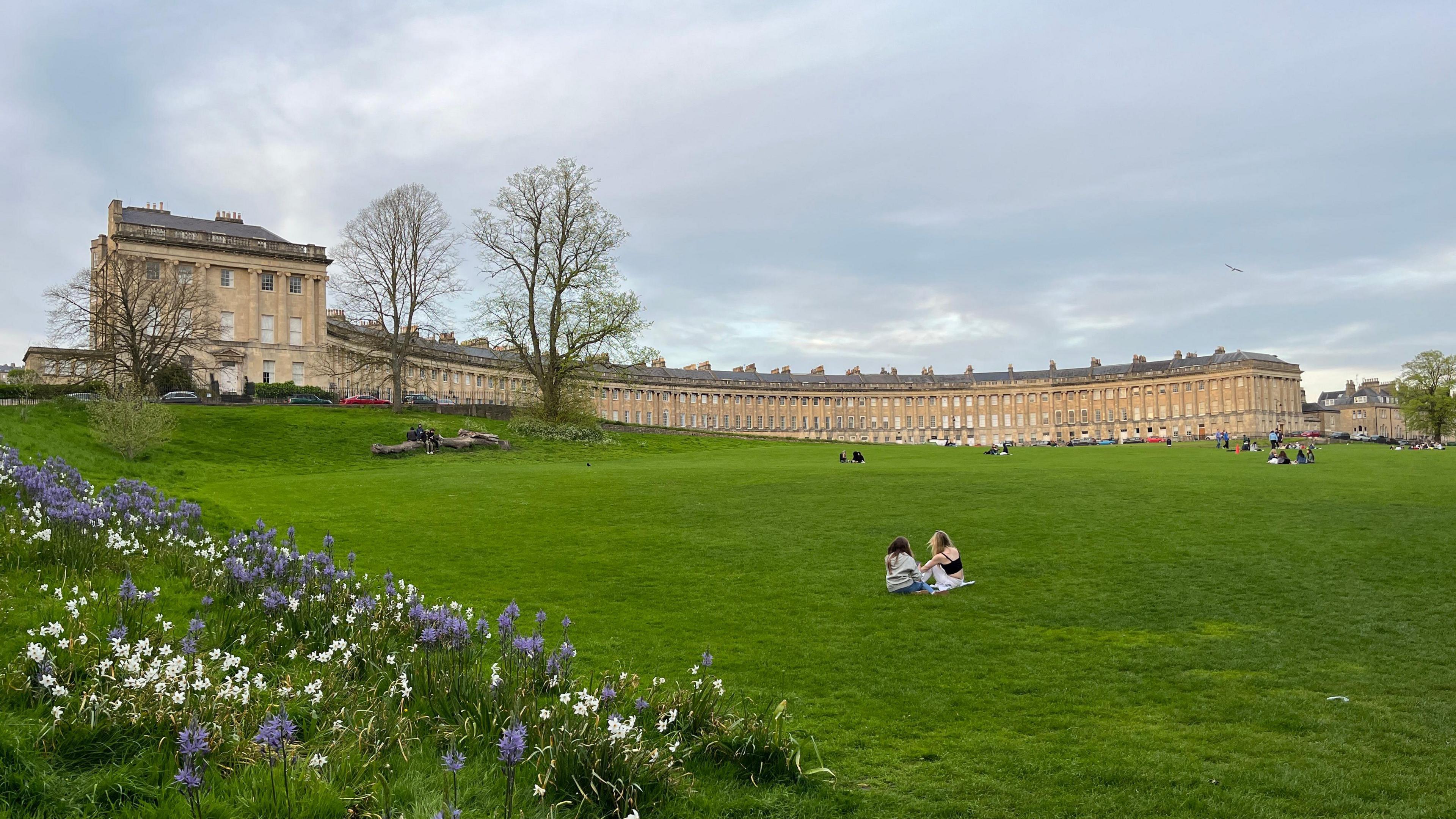 Bath's Royal Crescent seen from the green down the hill. There are people sitting on the green and flowers are blooming in the foreground. The crescent sits on top of the hill.