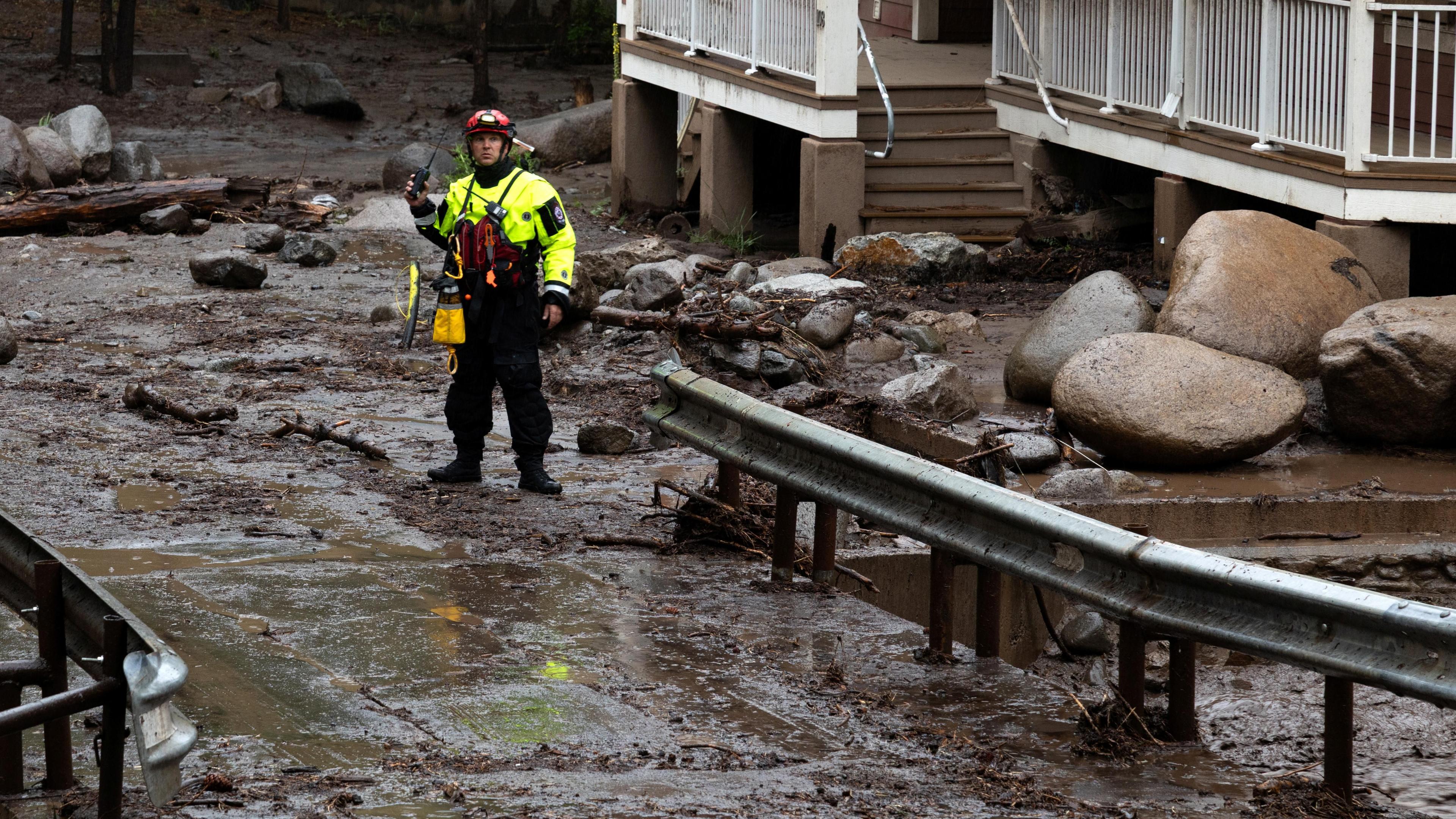 A male first responder dressed in neon yellow PPE surveys debris and mud covering a roadway in Ruidoso, New Mexico.