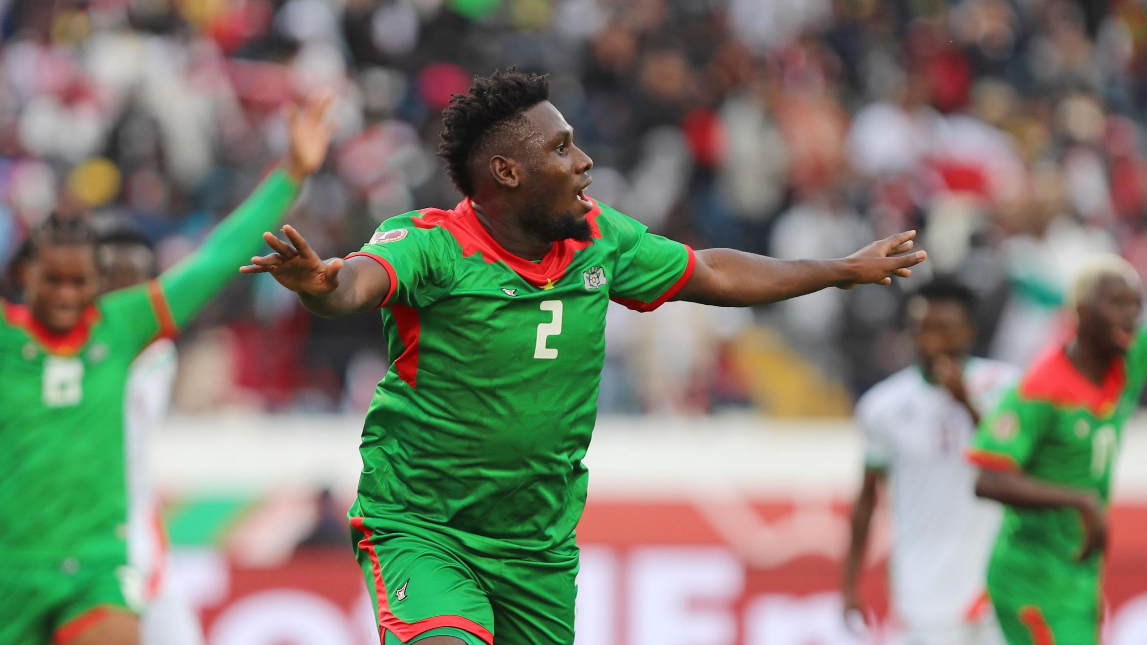 Lassina Traore, wearing a green Burkina Faso kit with red trim, stretches his arms out in celebration