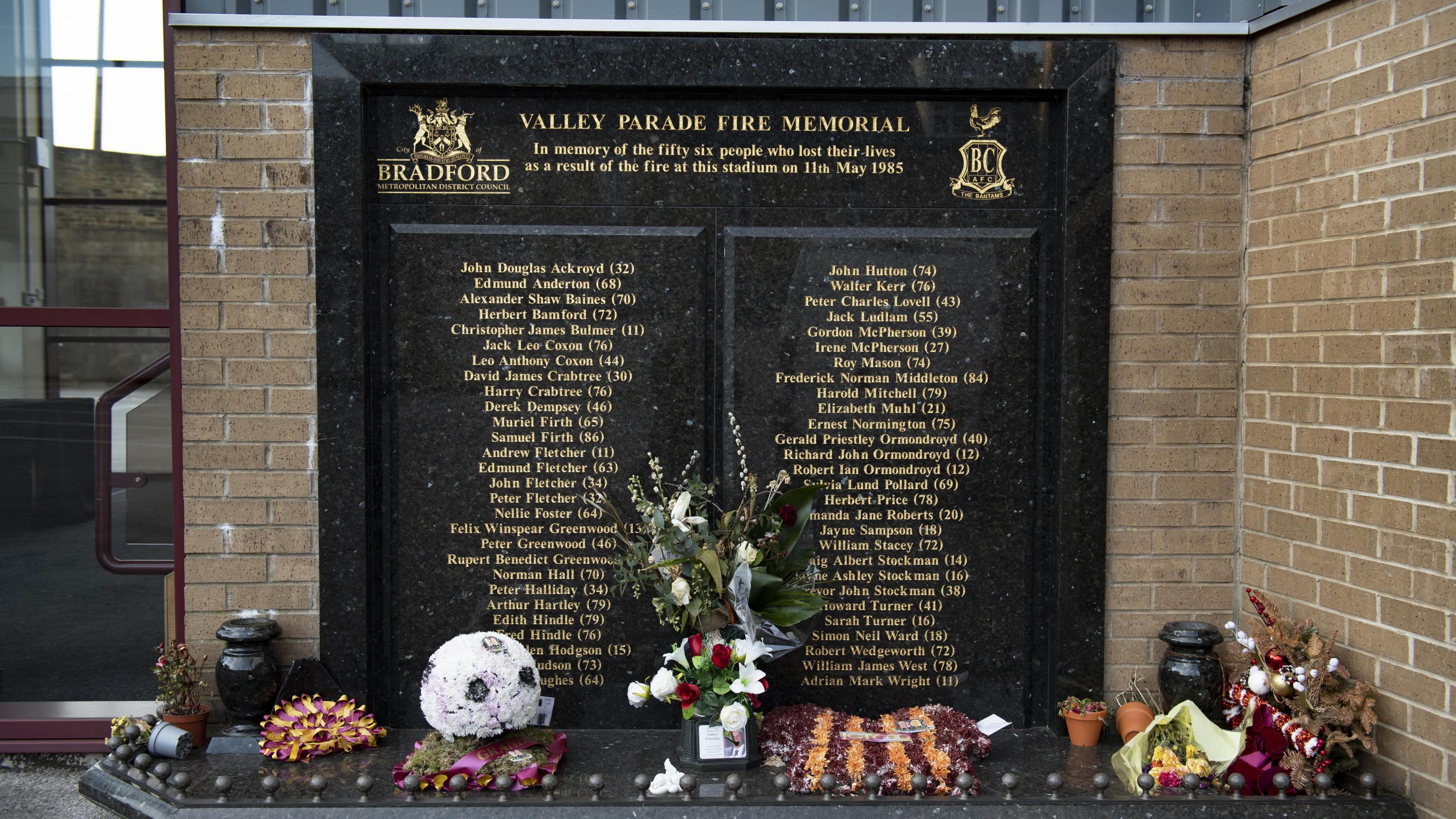 A stone memorial with floral tributes laid at the foot of it. A list of 56 names, 28 on either side, are engraved on it, with their ages next to them.