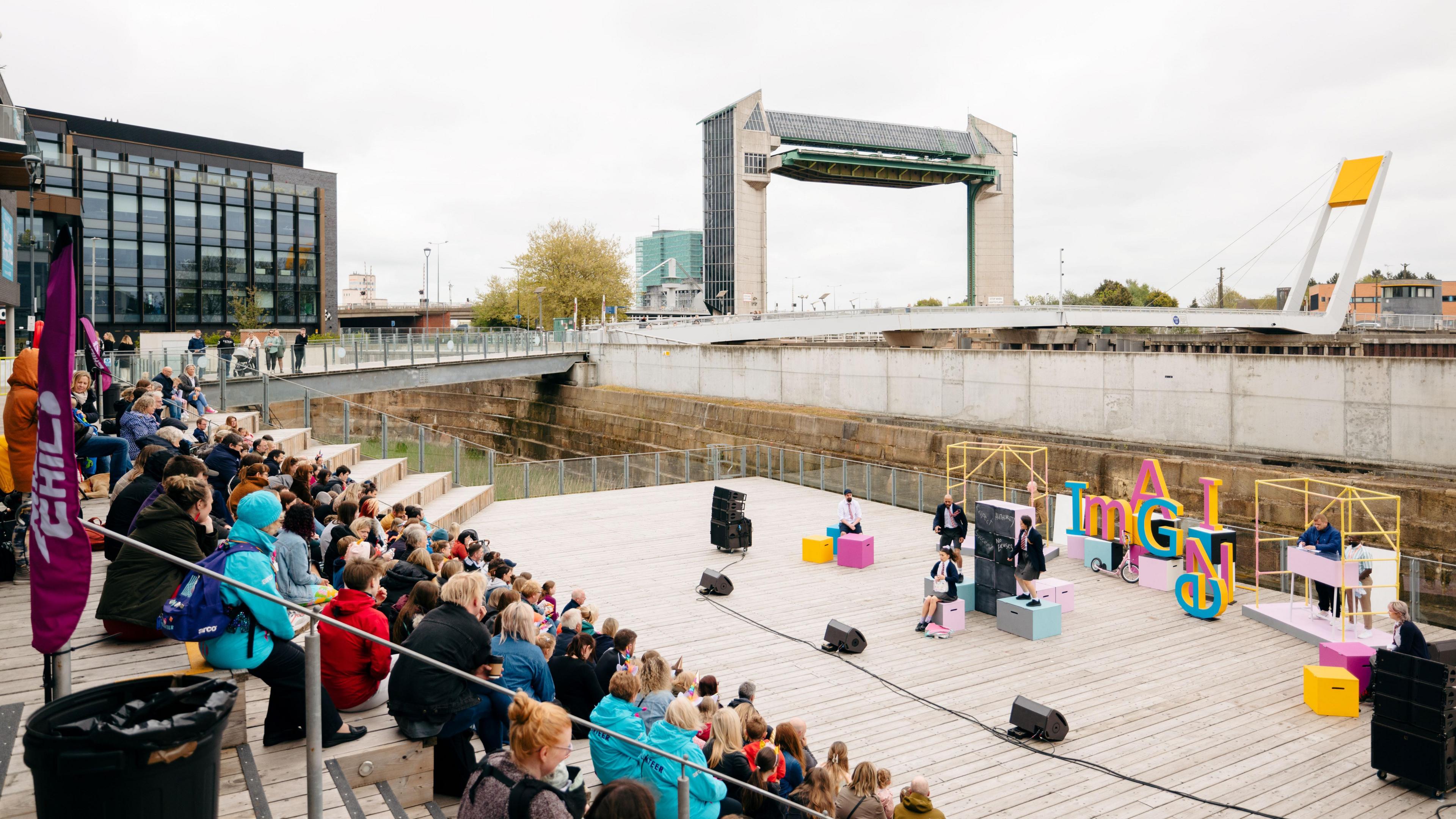 Crowds of people sat on steps watching a play on a wooden stage by a river in Hull. The sky is overcast.