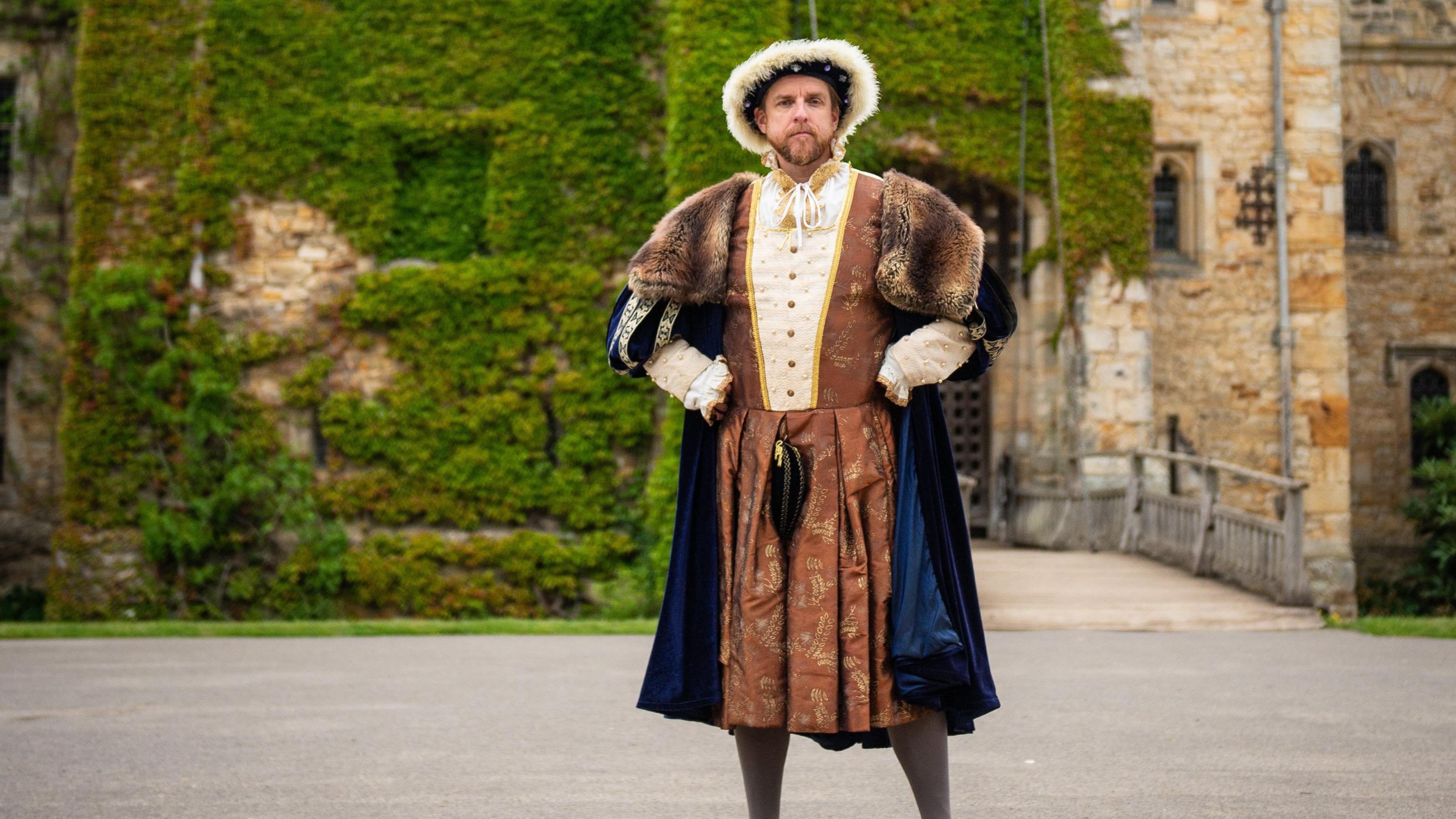 Mark Goldthorp dressed in gold and blue robes of Henry VIII posing with hands on hips outside Hever Castle