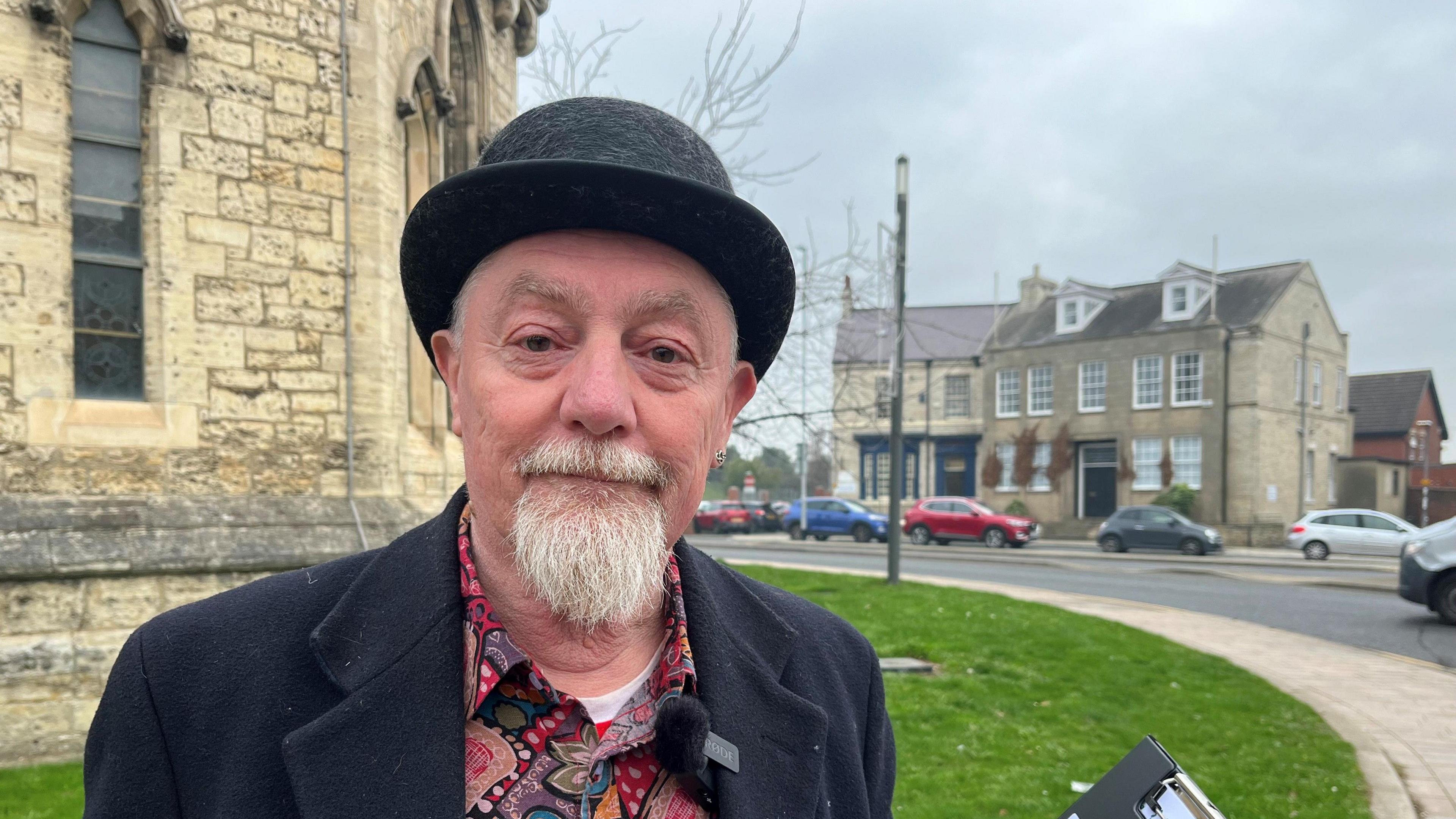 Petitioner Denis Lindridge outside where the tree has been positioned for years on Church Street, behind a church next to a busy road. He is wearing a bowler hat and colourful shirt.