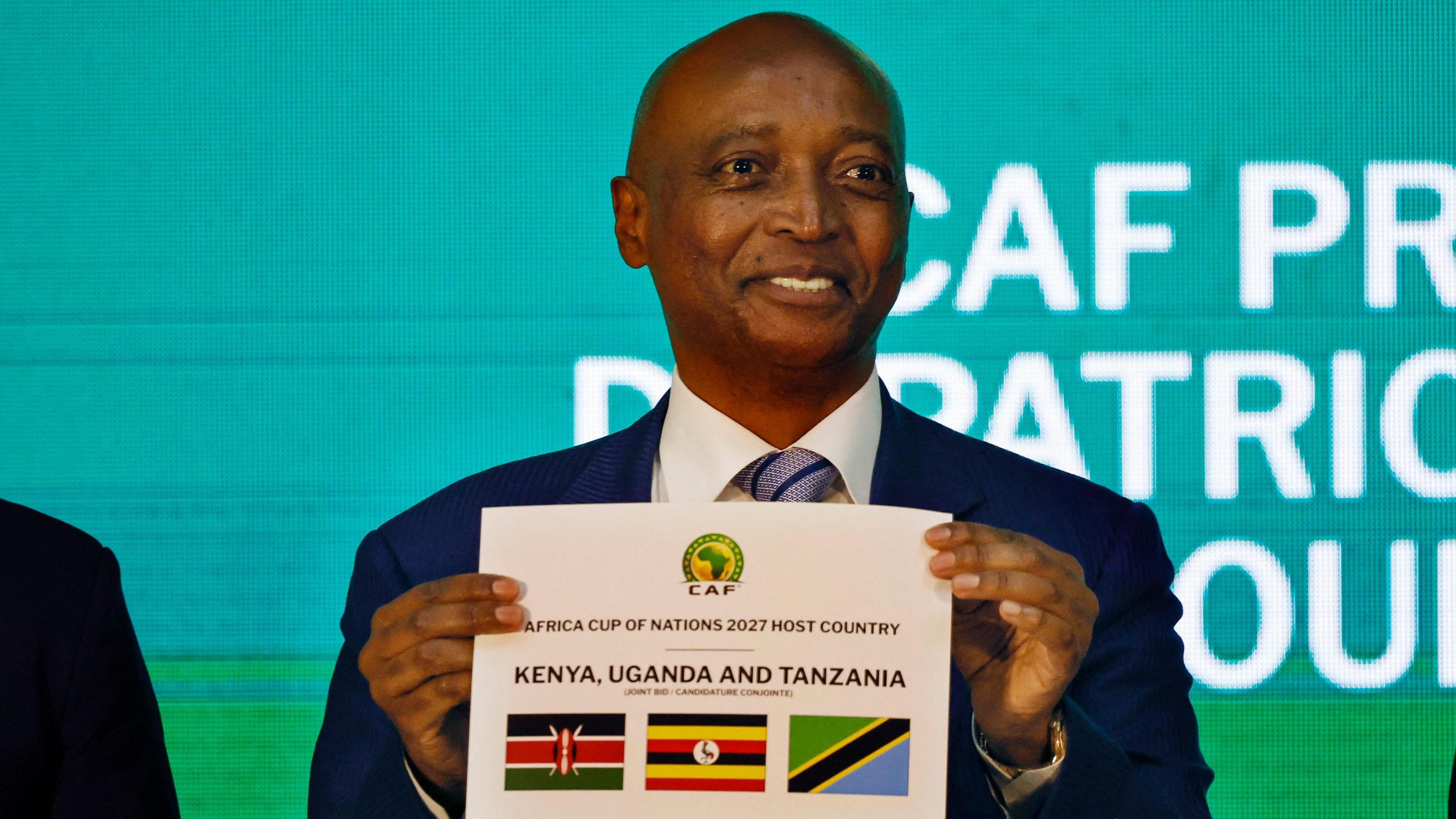 Patrice Motsepe, wearing a dark blue suit and patterned tie, holds up a piece of paper with flags detailing Kenya, Uganda and Tanzania as the three co-hosts of the 2027 Africa Cup of Nations