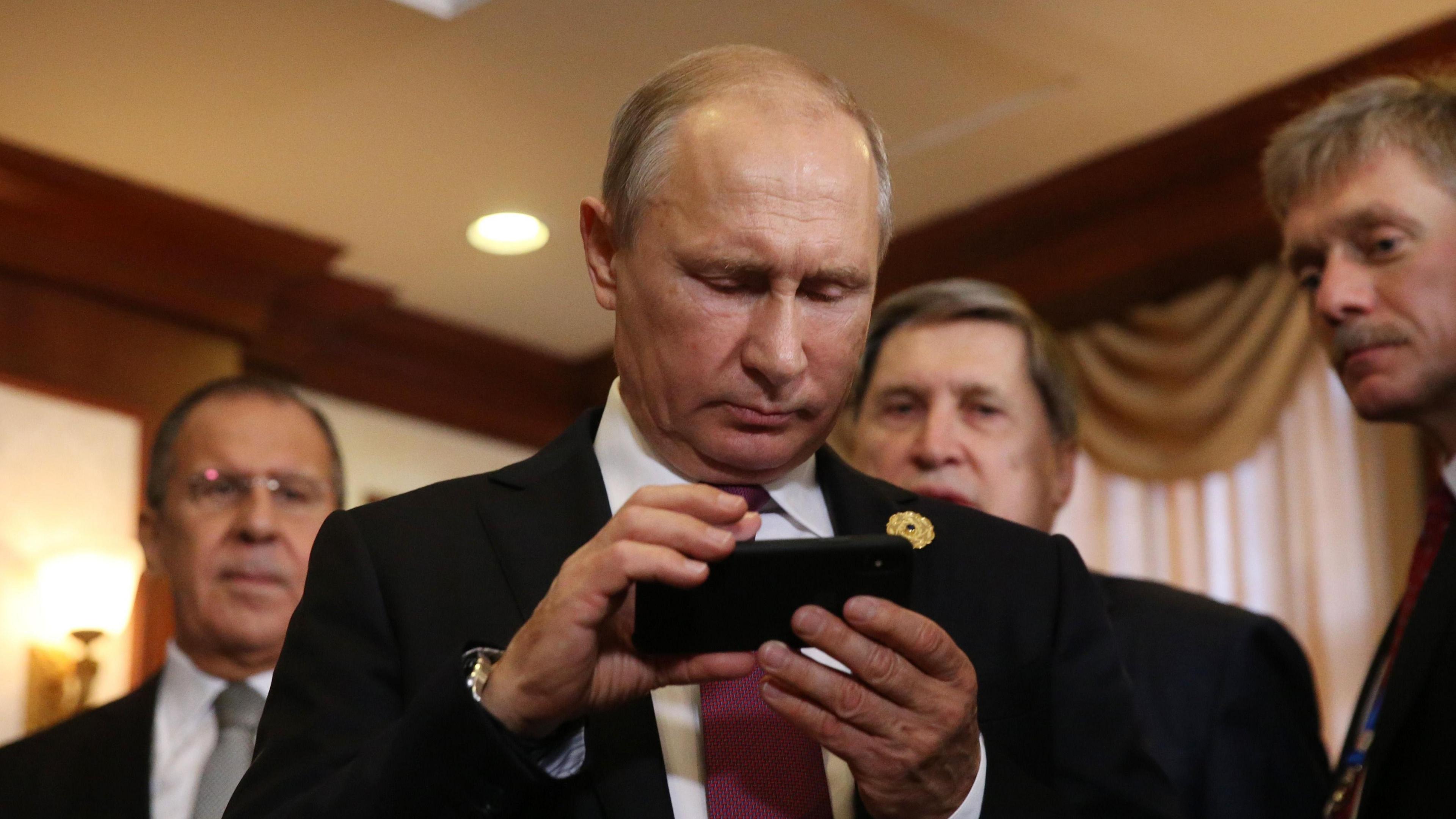 Photograph showing Russian President Vladimir Putin holding an iPhone, with Russian officials standing behind him in a formal setting.