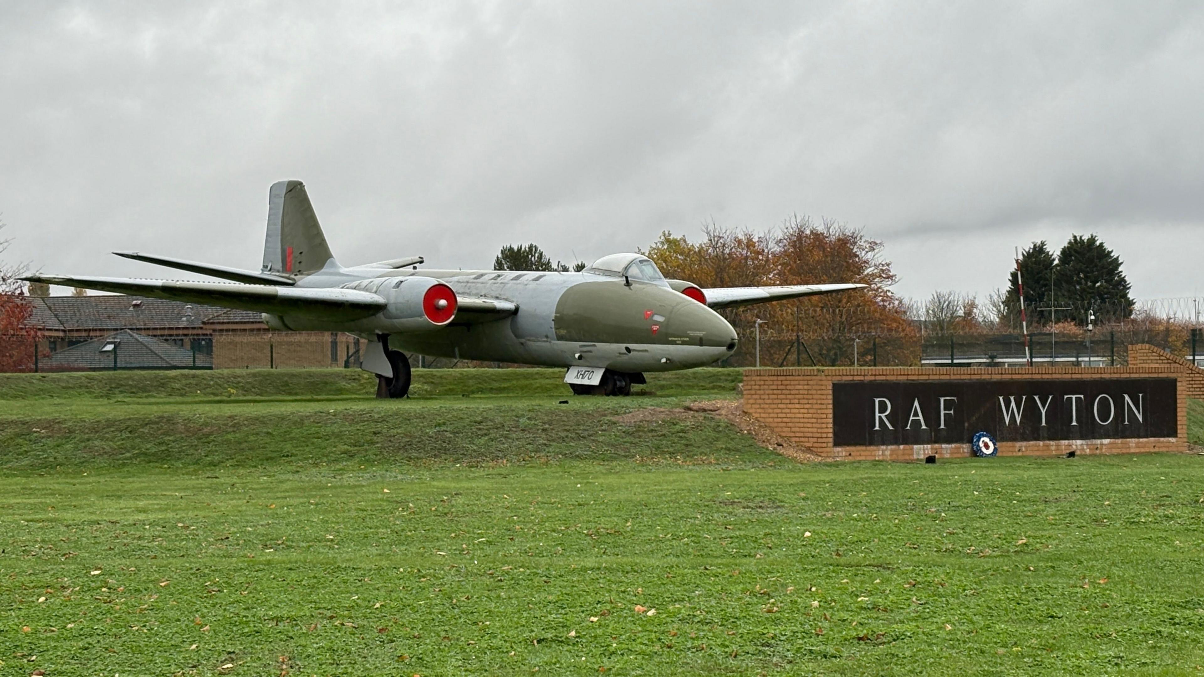 An old-looking jet plane outside RAF Wyton. The plane is grey and khaki green, and is sat on top of a raised, grassy platform. In front of its nose cone, on the right, is a name plate bearing "RAF Wyton", set into a low brick wall. There is an expanse of grass in the foreground and grey skies above the plane. In the background are autumnal-looking trees, whose leaves have turned orange.