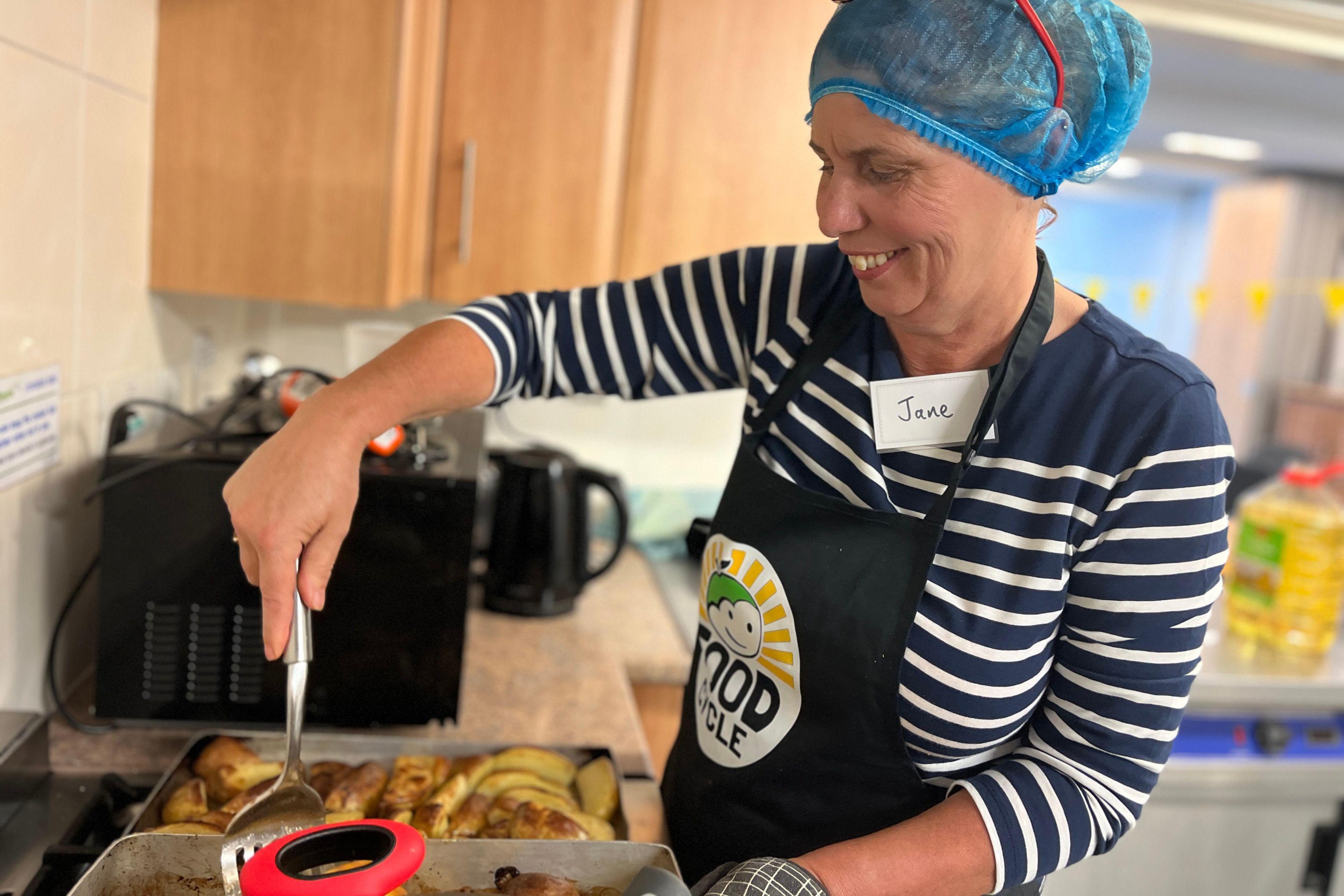 Volunteer cook Jane Hunt turns over some potato wedges in a pan. She is wearing a branded FoodCycle apron and a blue hair net