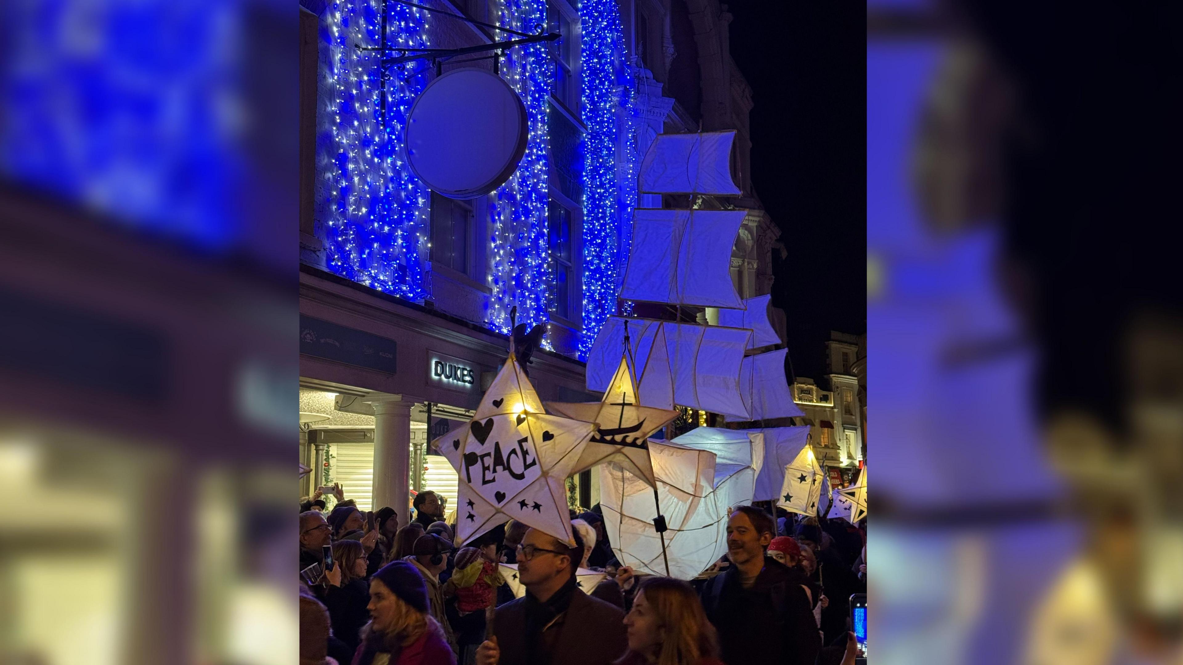 Lanterns are held in the air above a crowd parading through the street. The first lantern is in the shape of a star and has the word peace on it with stars and hearts also on it. Behind there is one in the shape of a large sailing boat, all are passing a shop building with blue fairy lights strung over its first floor level.