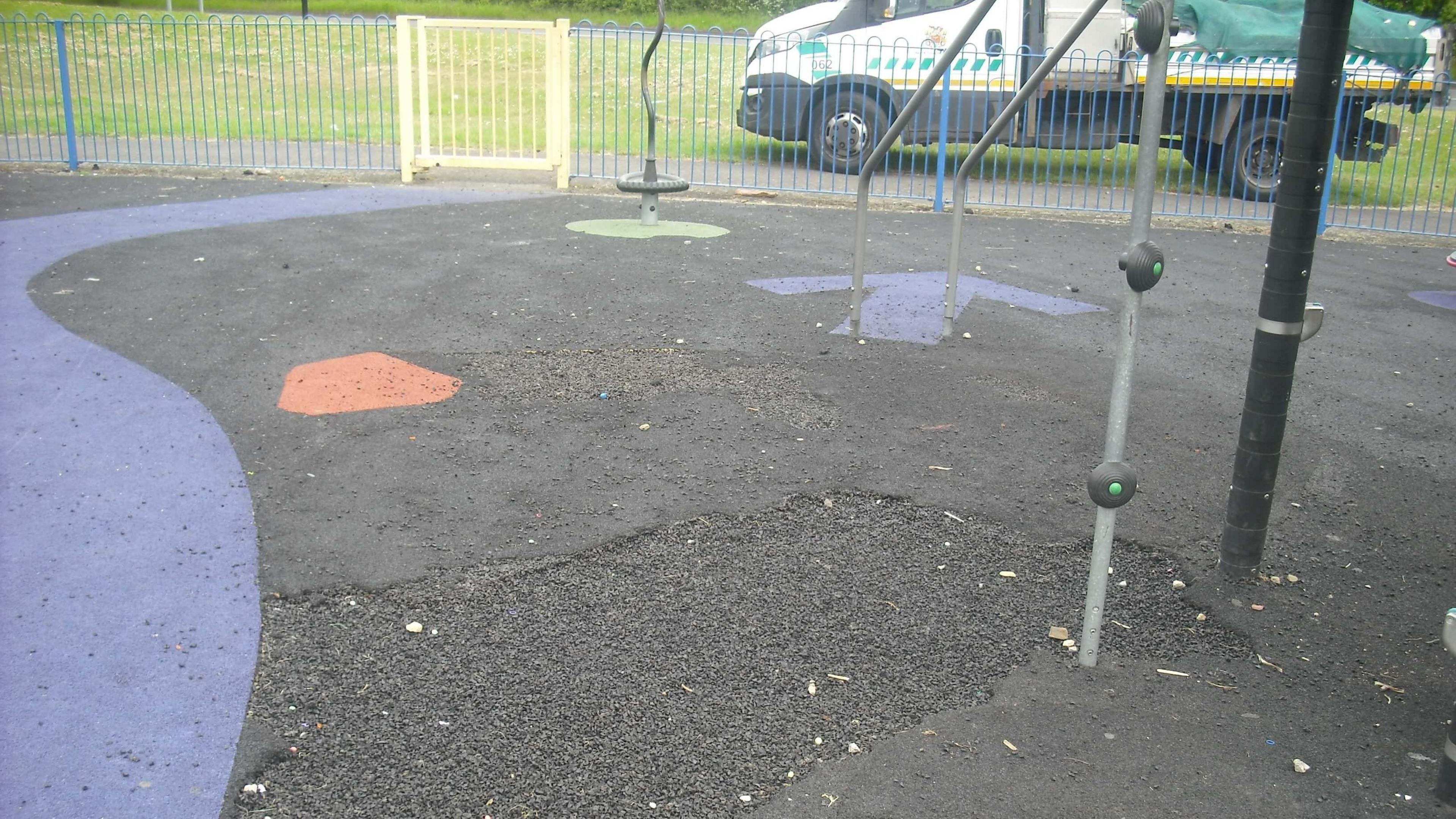 Black rubber surfacing with blue and orange spots next to play equipment. A large hole is in the floor.