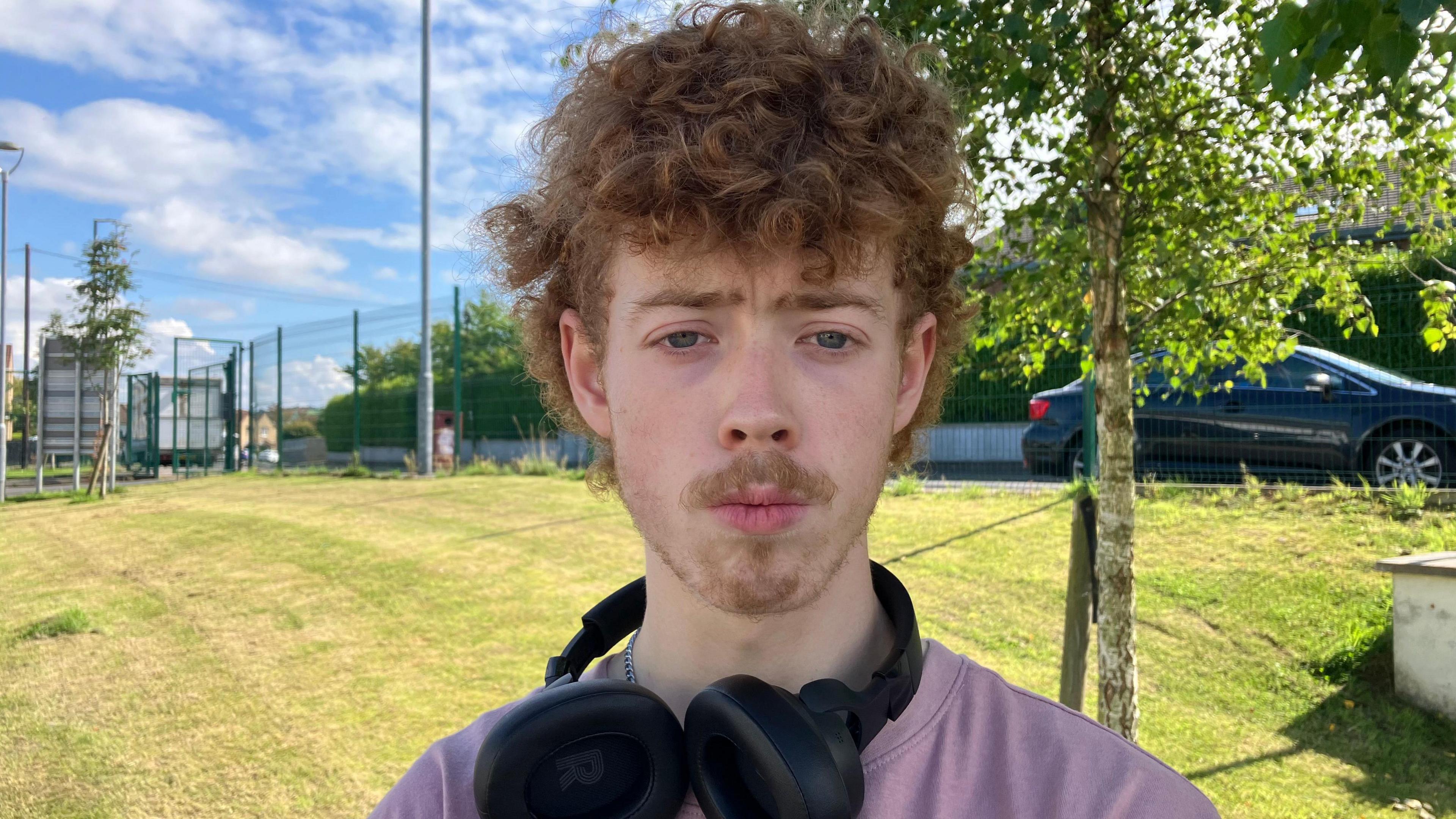 A young man standing outside on green grass with a tree behind him. He wears a light purple tshirt and has headphones around his neck.