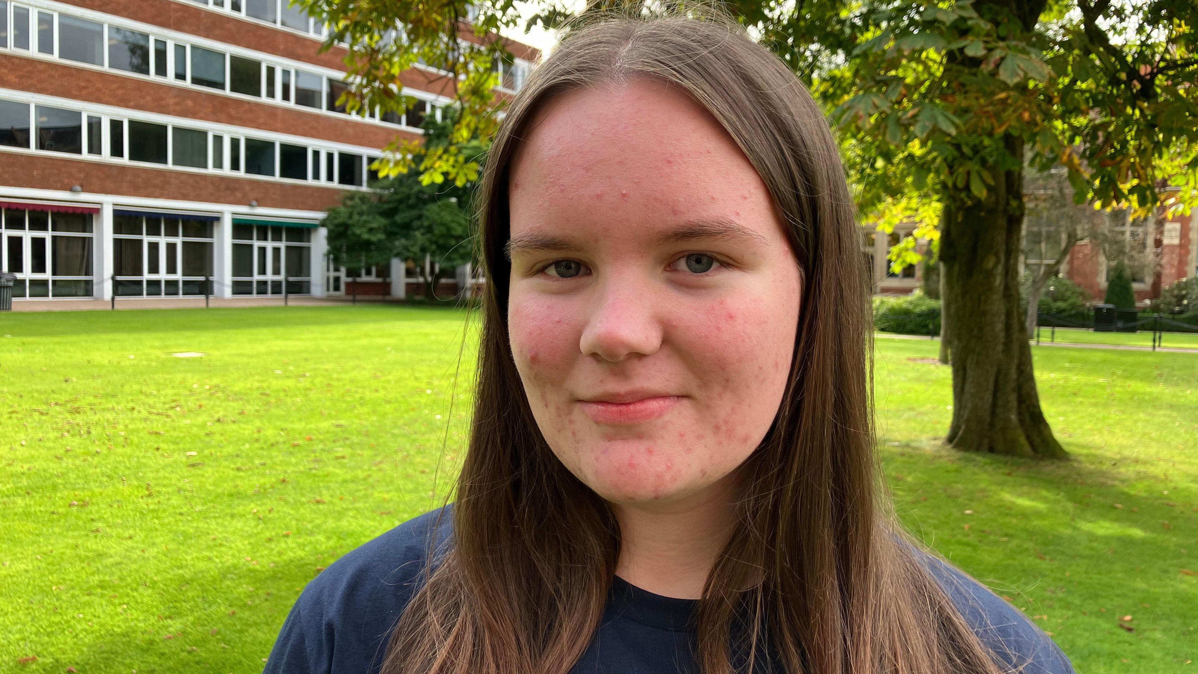 Maddison is standing in front of a three story building with lots of windows and red bricks in between the windows. There are trees behind her.

She has brown hair in a middle parting. She is looking at the camera and appears to have blue eyes.

She is wearing a blue long sleeved t-shirt. 