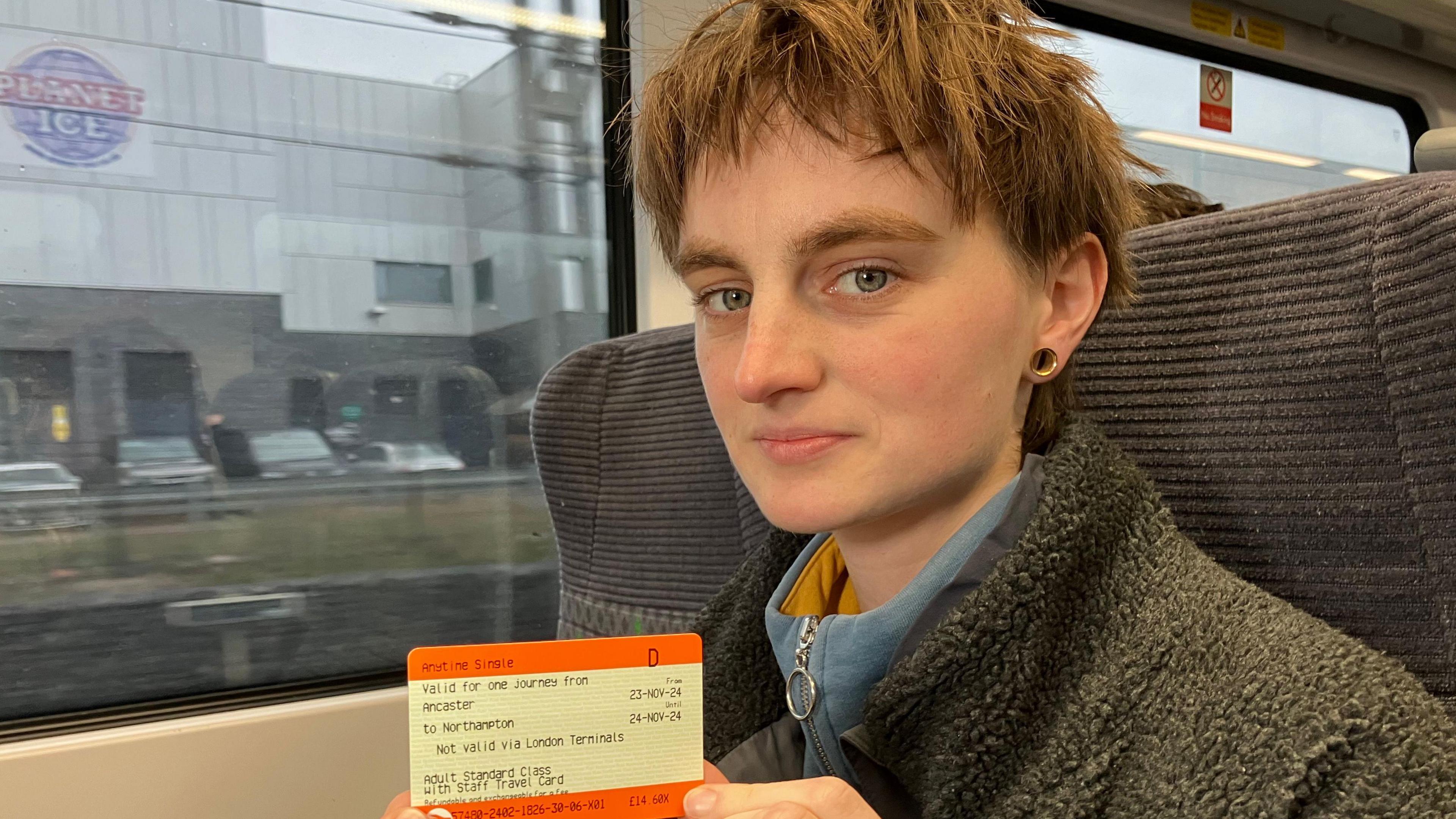 Mags Luckhurst wearing a grey fleece over a blue zipped up jacket and holding up a train ticket. She has short light brown hair and is sitting in a train