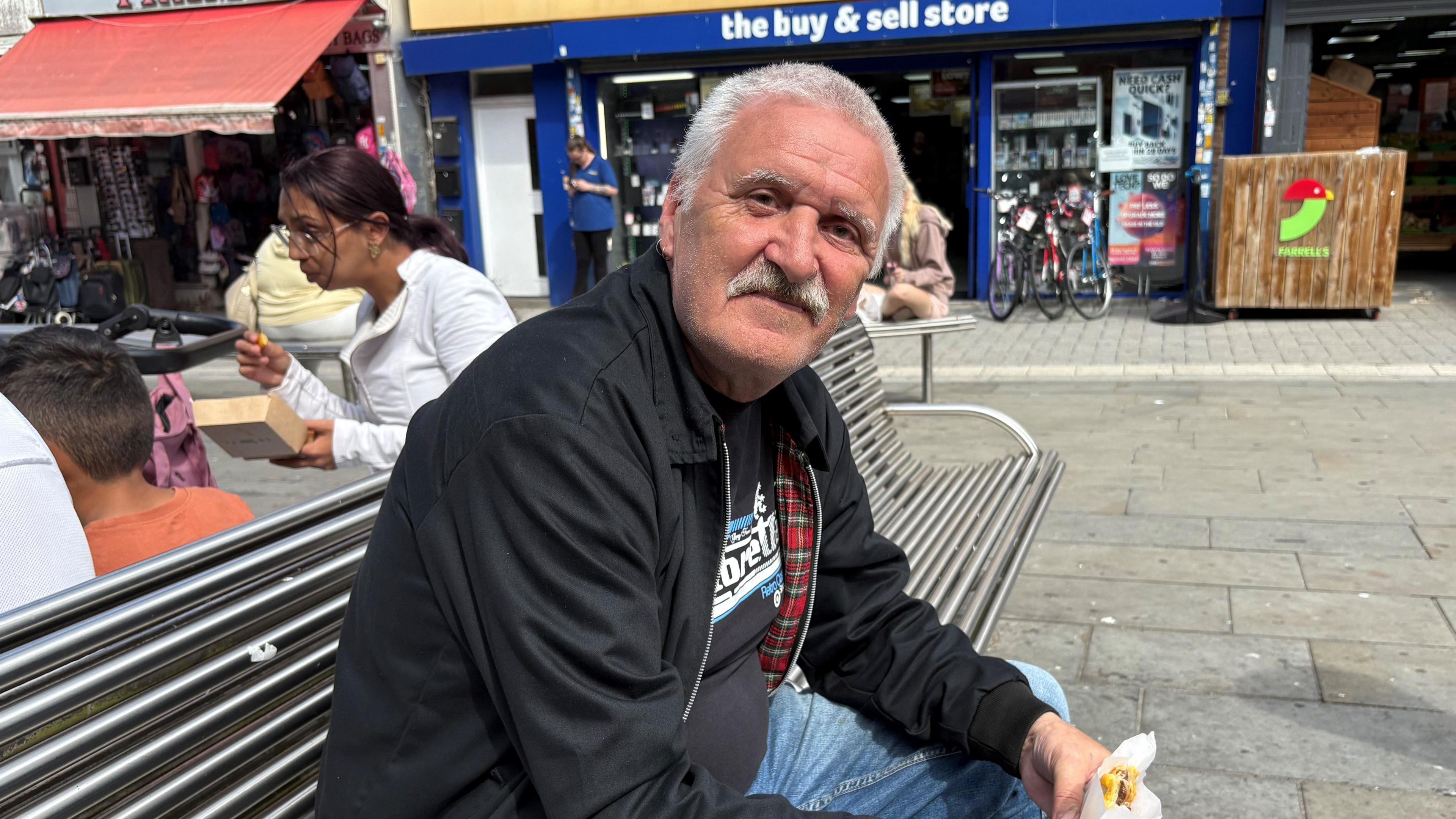 A man with a moustache and black Harrington jacket sits smiling on a bench while eating a sandwich.
