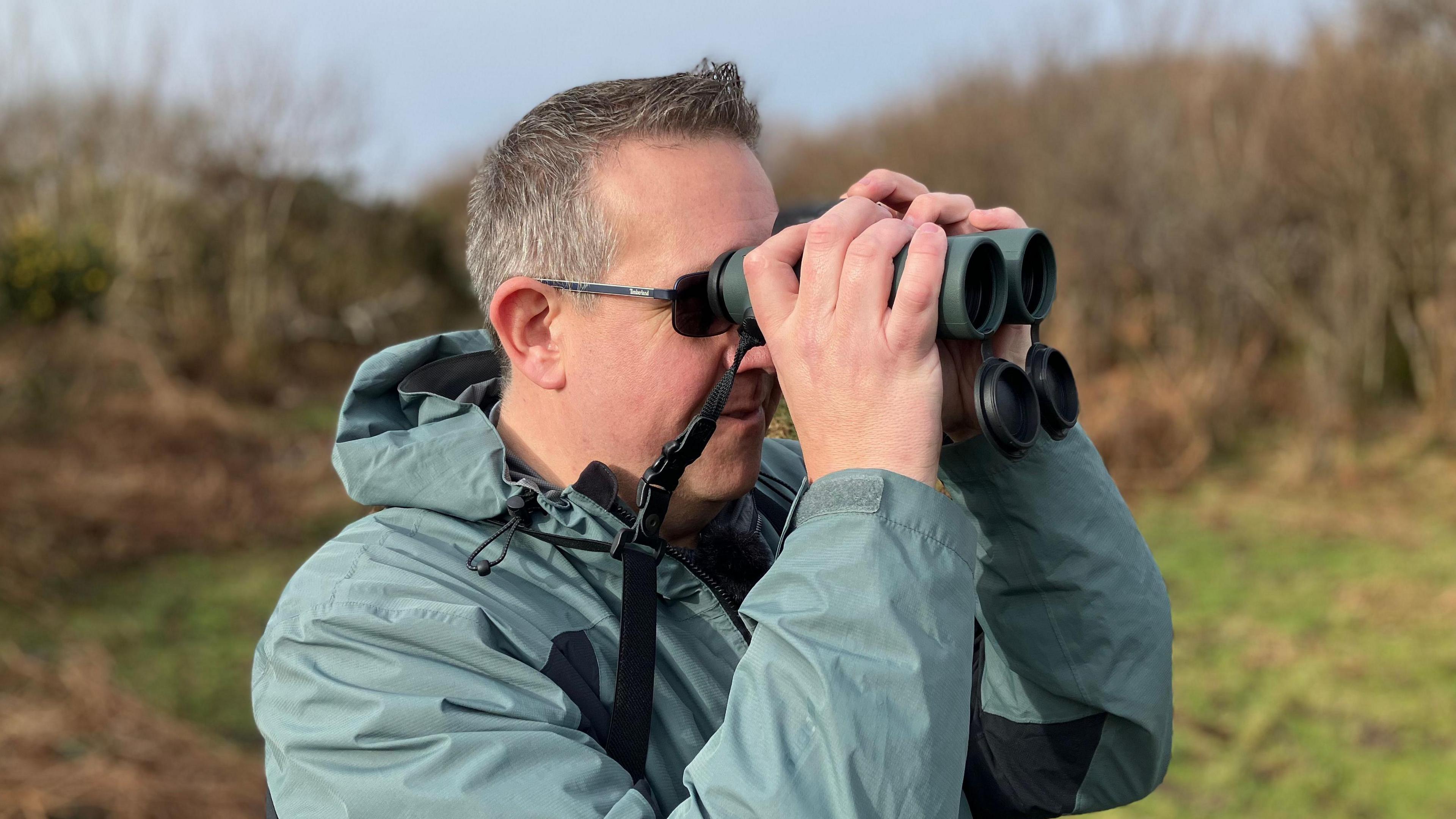 A birdwatcher is standing in the countryside, with moorland scrub behind him. He is wearing a green waterproof coat and looking through green binoculars. 