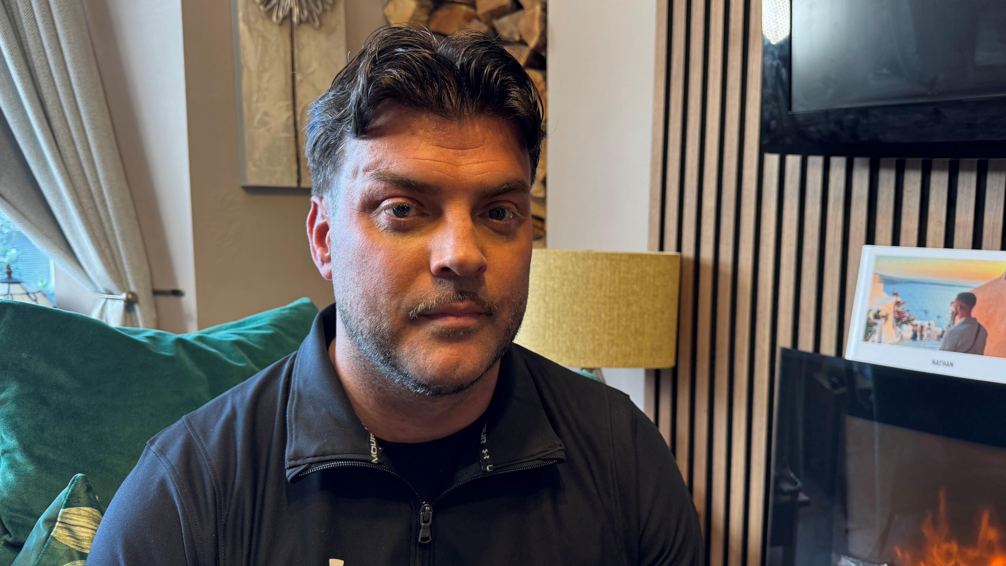 Lee has dark short hair and stubble, and is looking at the camera with a straight face. He is wearing a black sports jacket with a zip, and a green sofa cushion can be seen behind him. On the right hand side is a panelled wooden wall with a wood burner at the bottom, flames can be seen through the glass. Above, a black TV screen is mounted on the wall. 