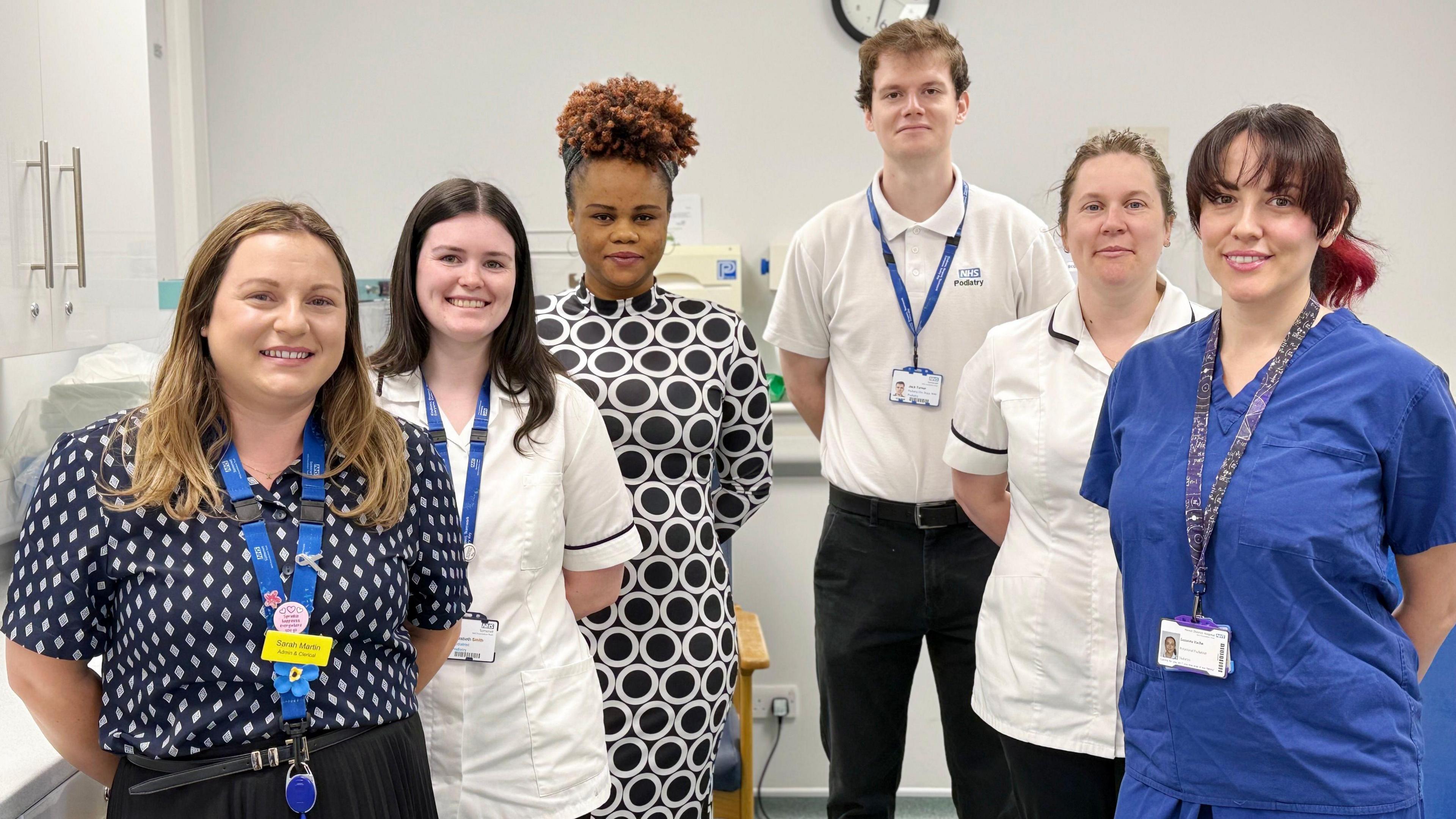 A group of six healthcare workers. Five of them are women and there is one man. They are all stood with their arms behind their backs, looking at the camera and smiling. 