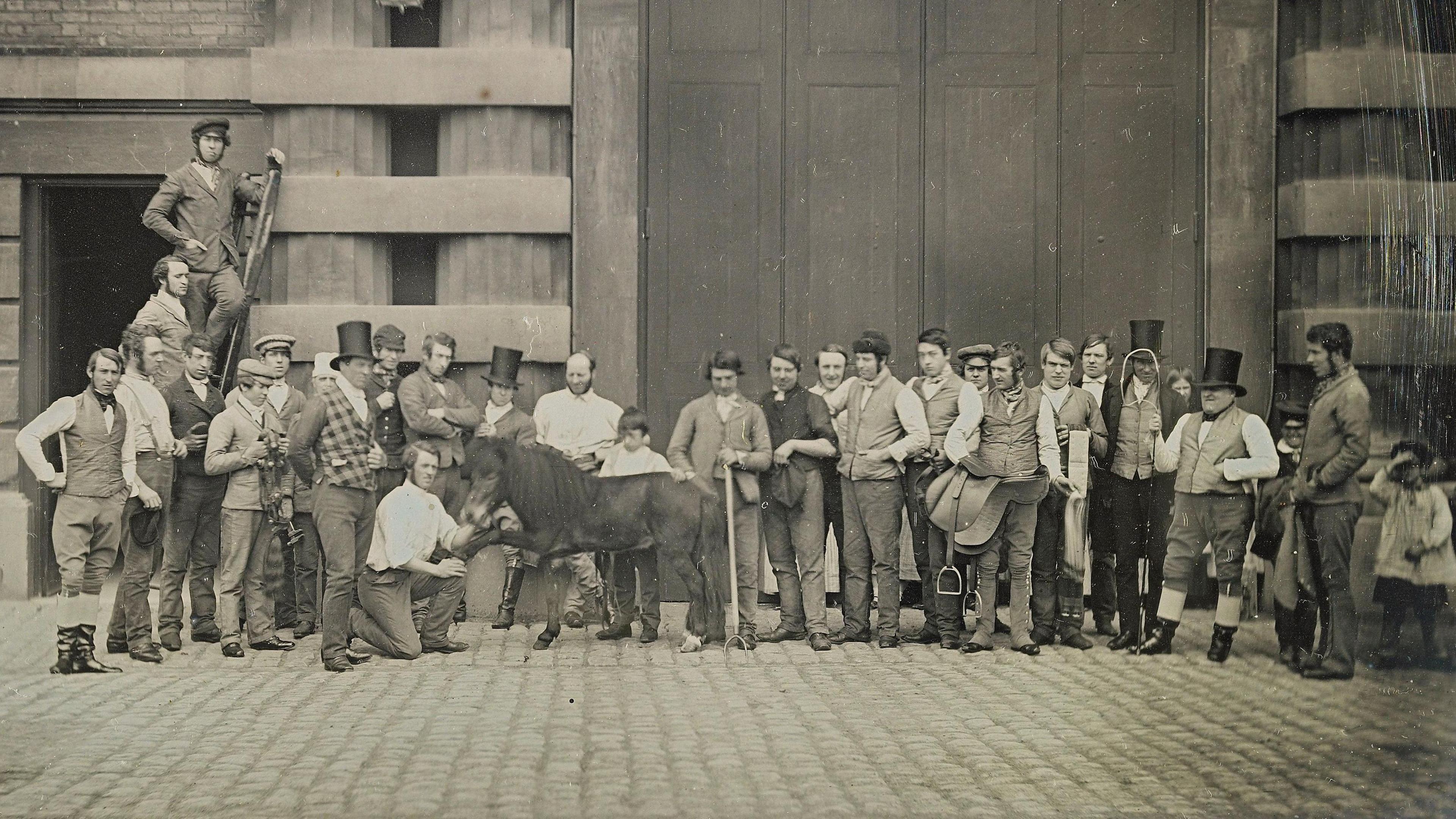 A 19th century image of large group of people standing in a line. One man is kneeling by a pony who is holding its hoof up.  