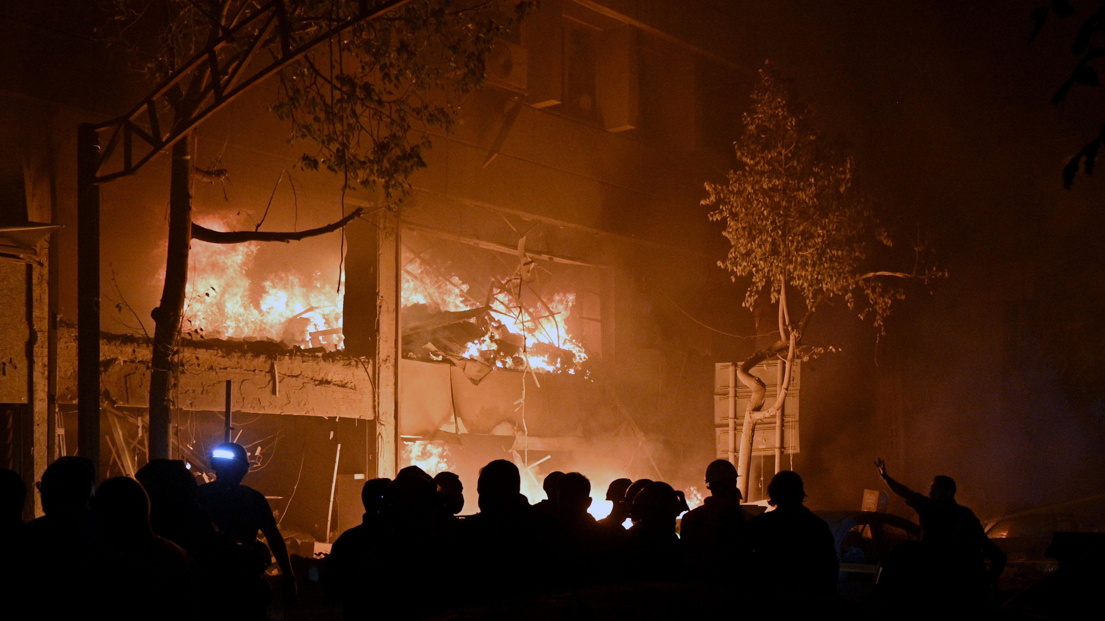 A building is in flames at night, with a crowd of firefighters and onlookers below