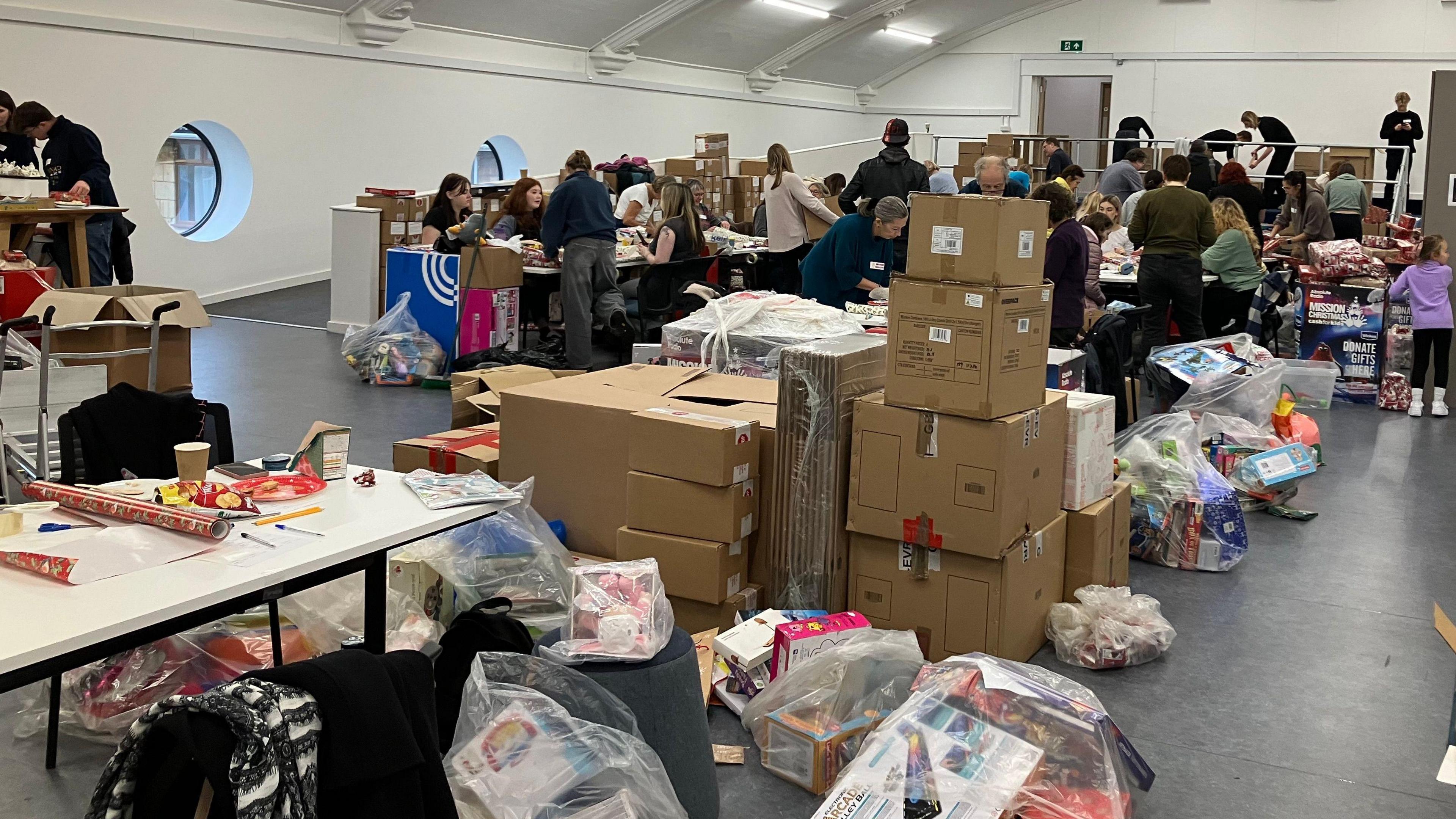 A large room is pictured with many boxes and bags of gifts visible on the bare floor. Dozens of people are also visible packing presents and talking to each other. The walls are white and have porthole-style windows