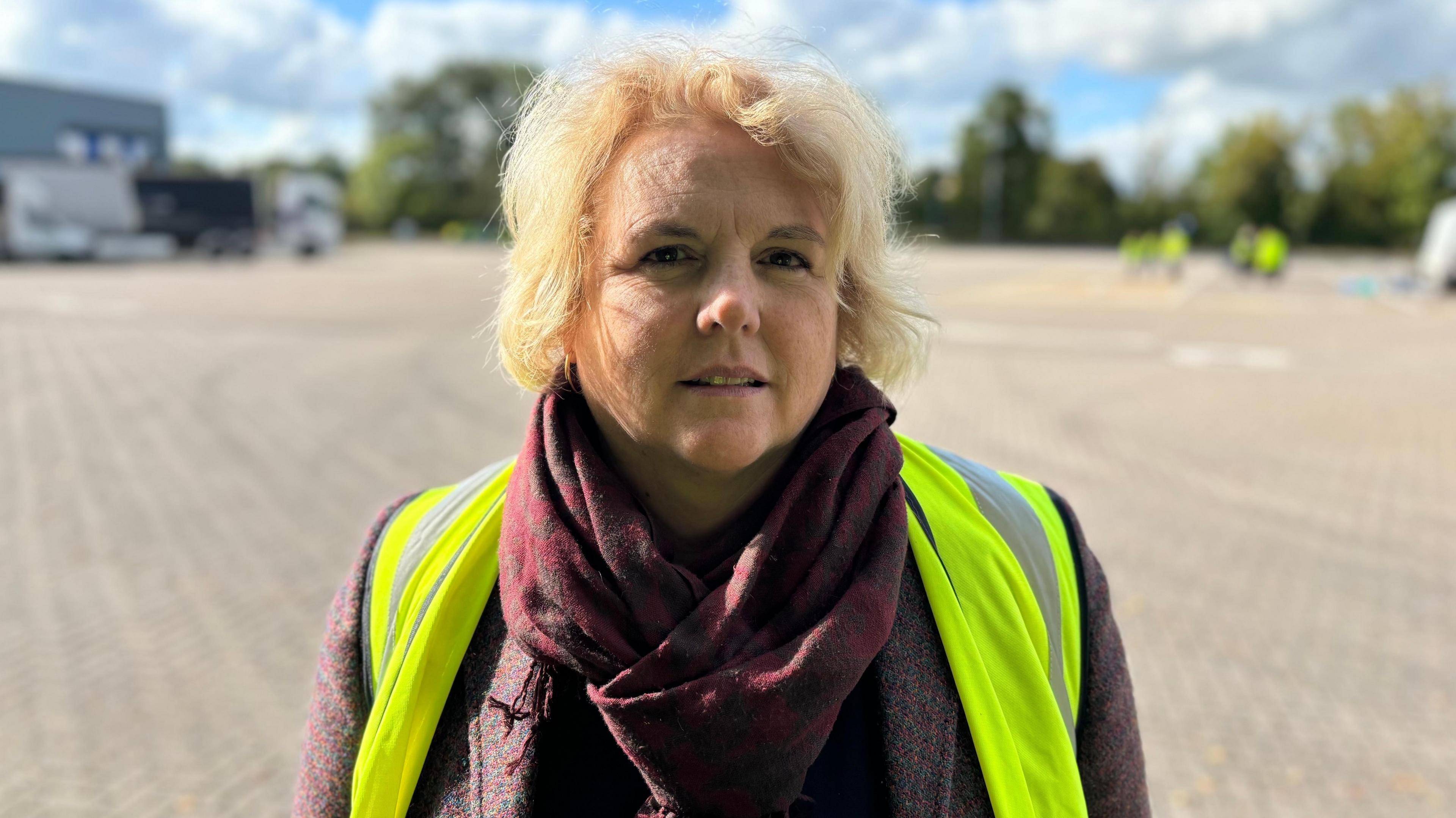 A woman with blonde hair wearing a hi-vis jacket and red scarf and coat, stands in sunshine.