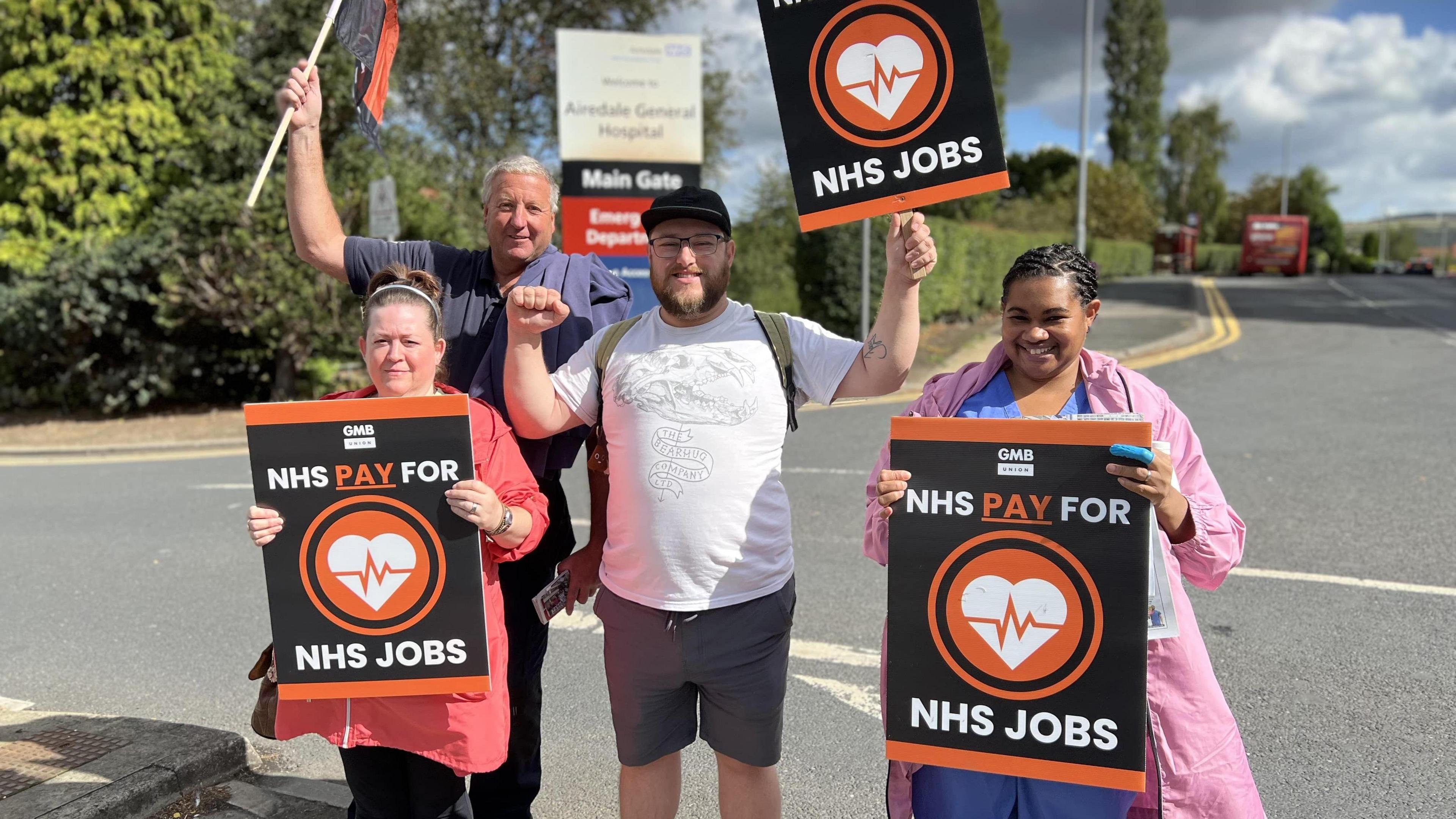 A group of four people stand on a roadside holding placards and flags saying "NHS Pay for NHS Jobs". One of them wears hospital scrubs. Behind them is the entrance and sign for Airedale General Hospital.