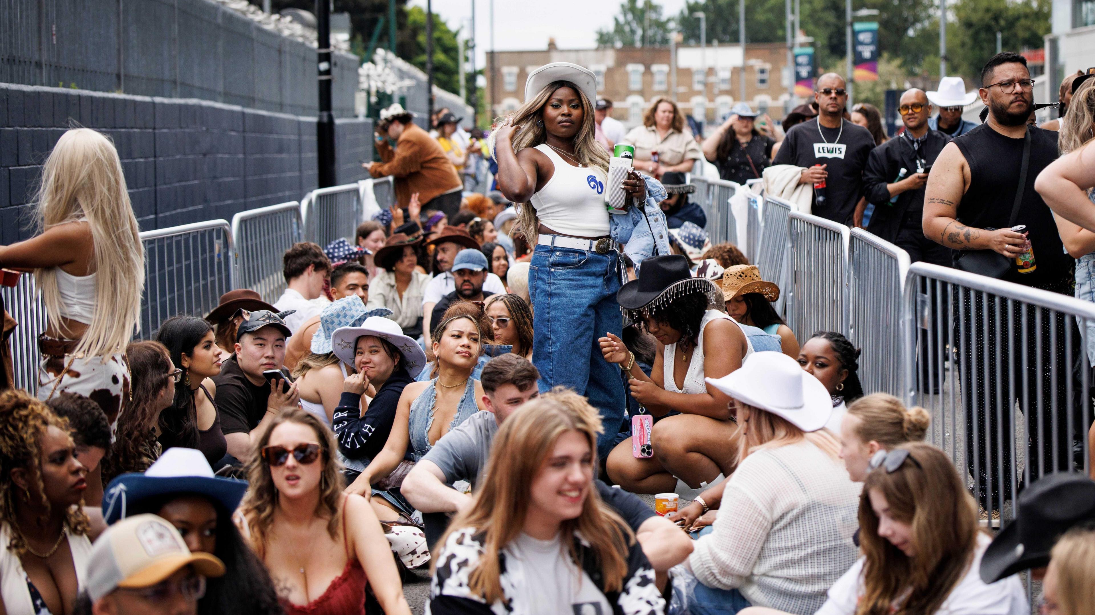 The queue waiting for the Beyoncé concert in Tottenham Hotspur Stadium. There are dozens of people sat on the ground between metal fencing. Others are walking past to their right.