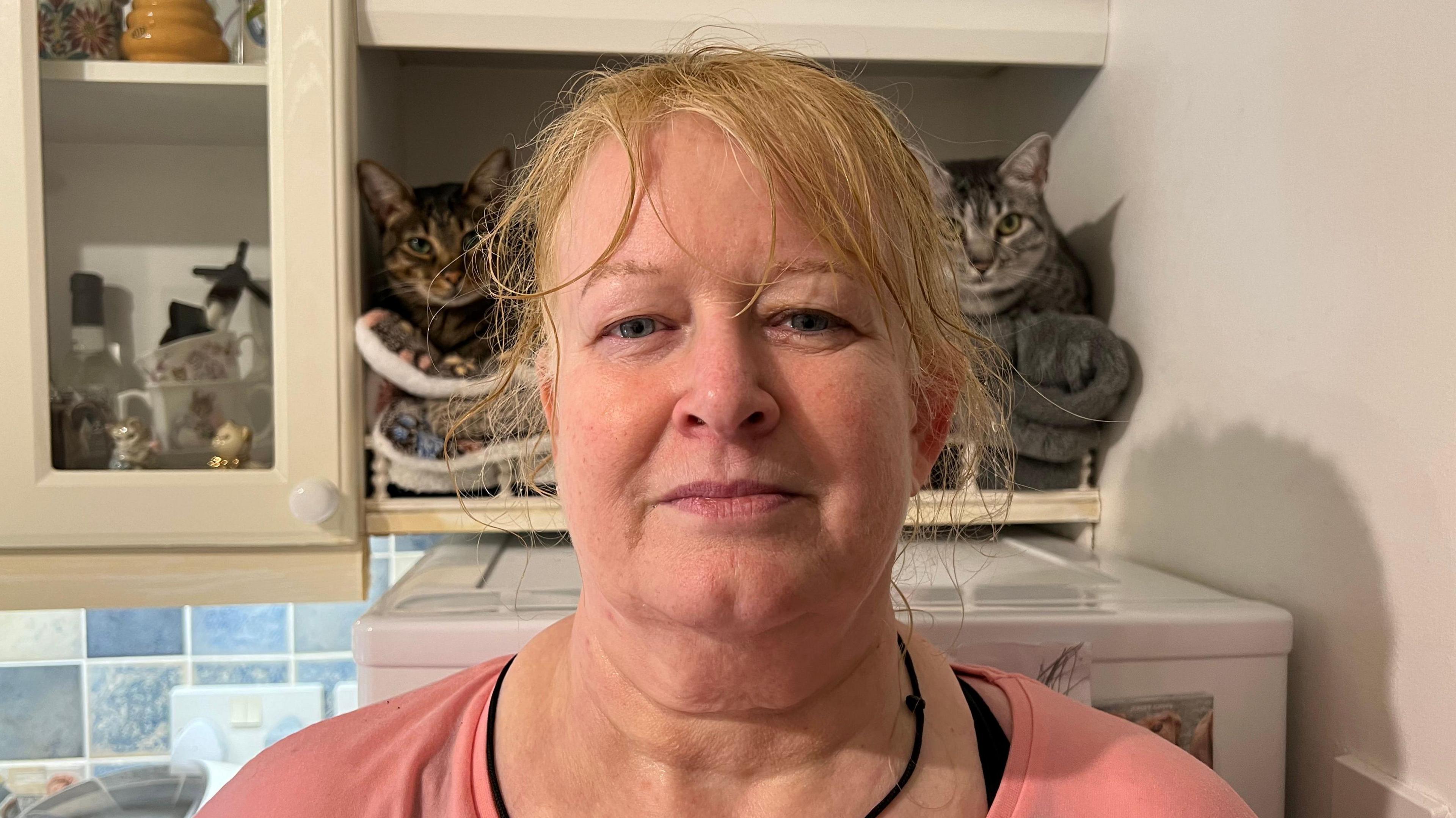 A woman with blonde hair across her forehead. She is looking into the camera and wearing a pink top with a black cord necklace. The background is a kitchen shelf with a brown cat looking at the camera to her left and a grey cat to her right.