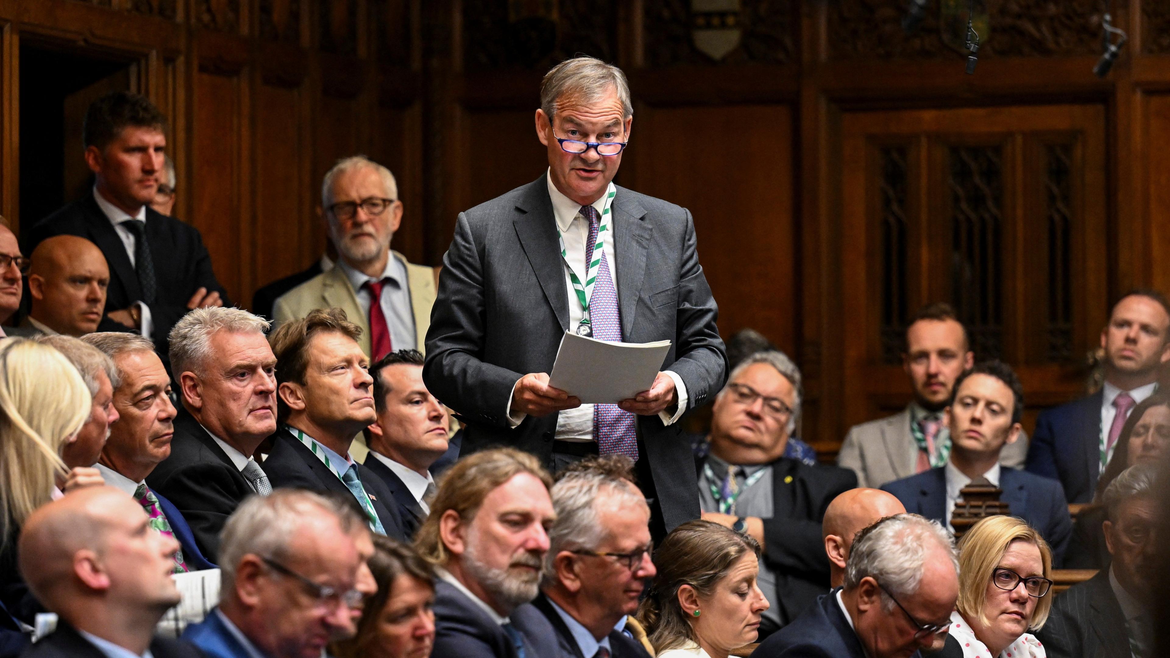 Rupert Lowe is standing surrounded by MPs in the House of Commons. He is holding paperwork and speaking, an is wearing a dark grey suit with a green striped lanyard around his neck