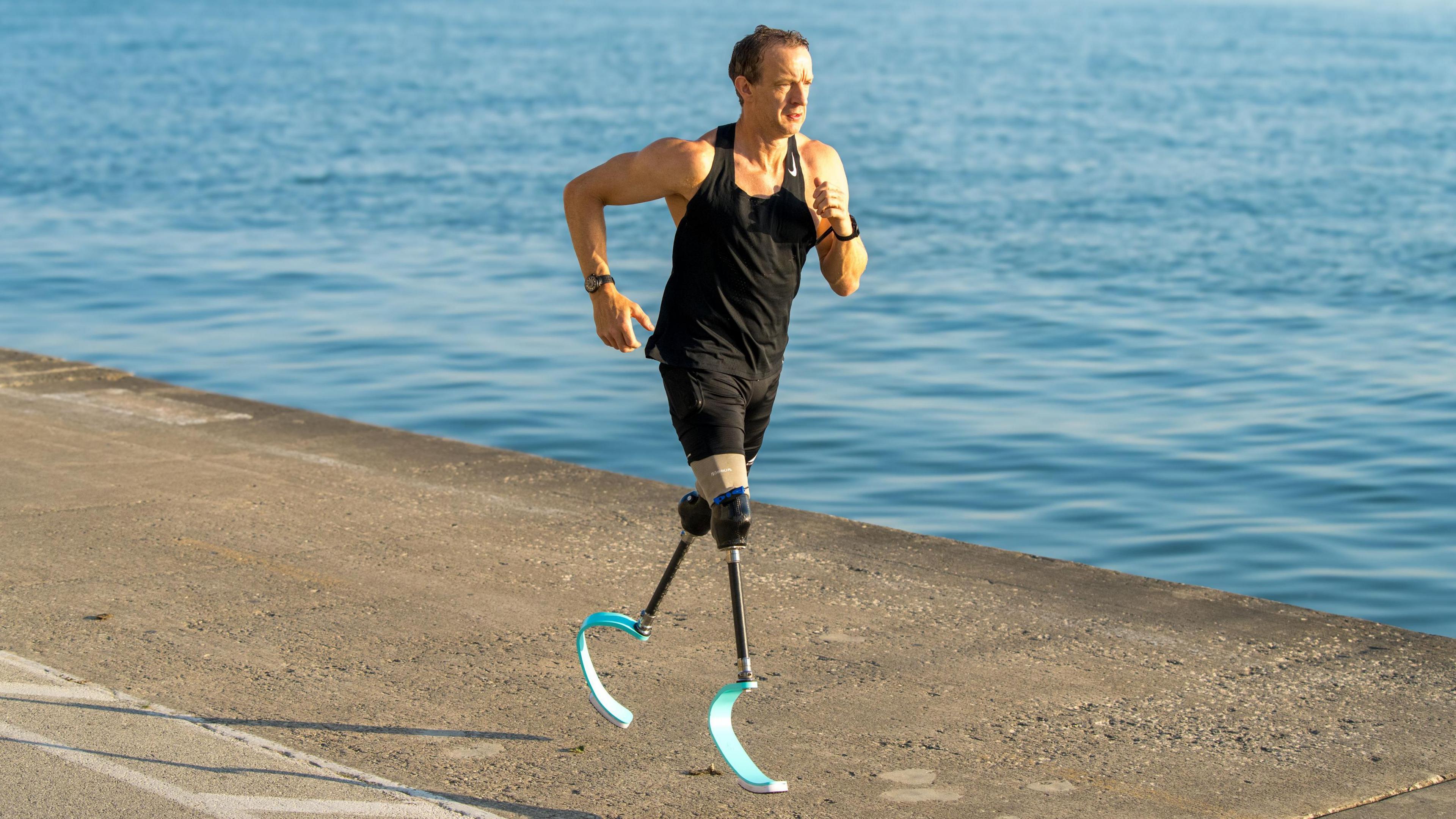 Richard is running along a tarmac surface alongside a body of clear blue water. He is wearing a black sleeveless vest and back shorts, he has short dark brown hair and has prosthetic lower limb blades.