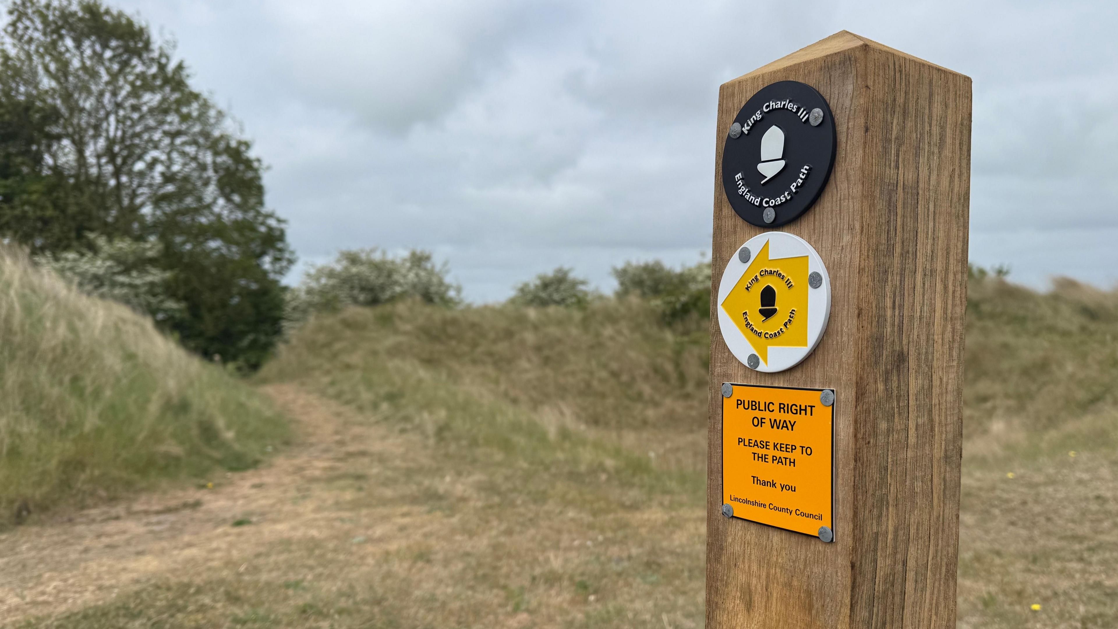 At the forefront of the photo on the right is a post, with three signs on. The top is a black circle, reading 'King Charles III England Coast Path.' The sign underneath is a white circle with a yellow arrow which is pointing to the left, also reading 'King Charles III England Coast Path.' The bottom line reads 'Public Right of Way.' In the background, to the left, is a path surrounded by grass and trees.